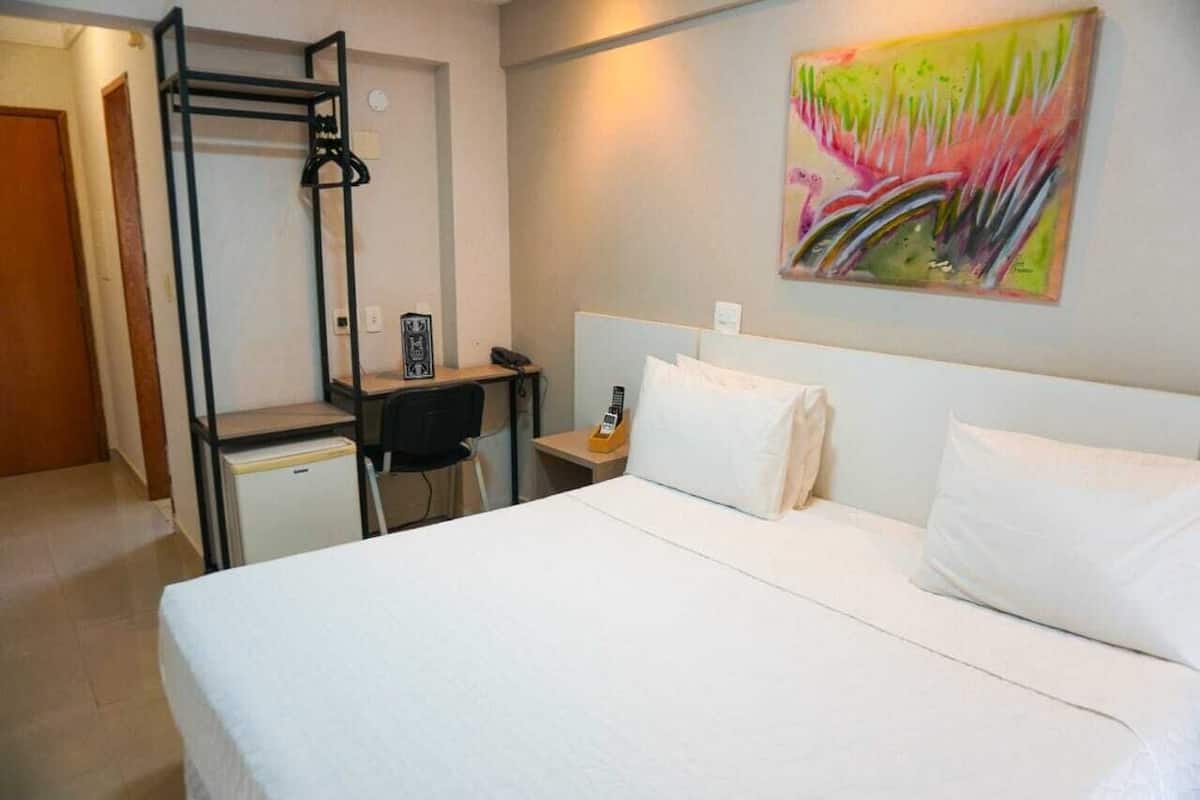 A hotel room with a bed, a desk with a chair, a small refrigerator, a telephone, and a painting on the wall.