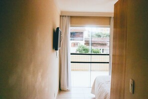 Ein Hotelzimmer mit Bett, einem an der Wand montierten Fernseher und einem Balkon mit Blick auf Gebäude und Grünflächen.