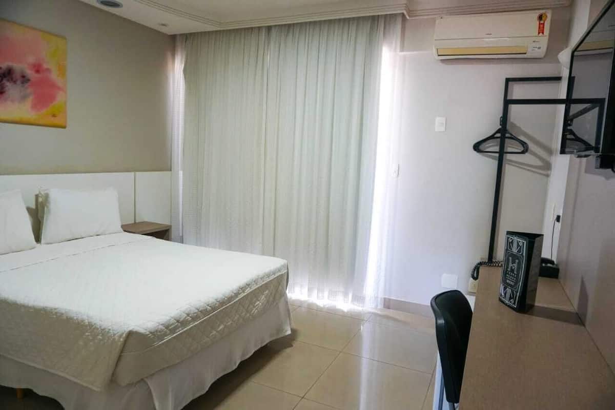 A hotel room with a bed, a chair, an air conditioning unit, and a coat rack.