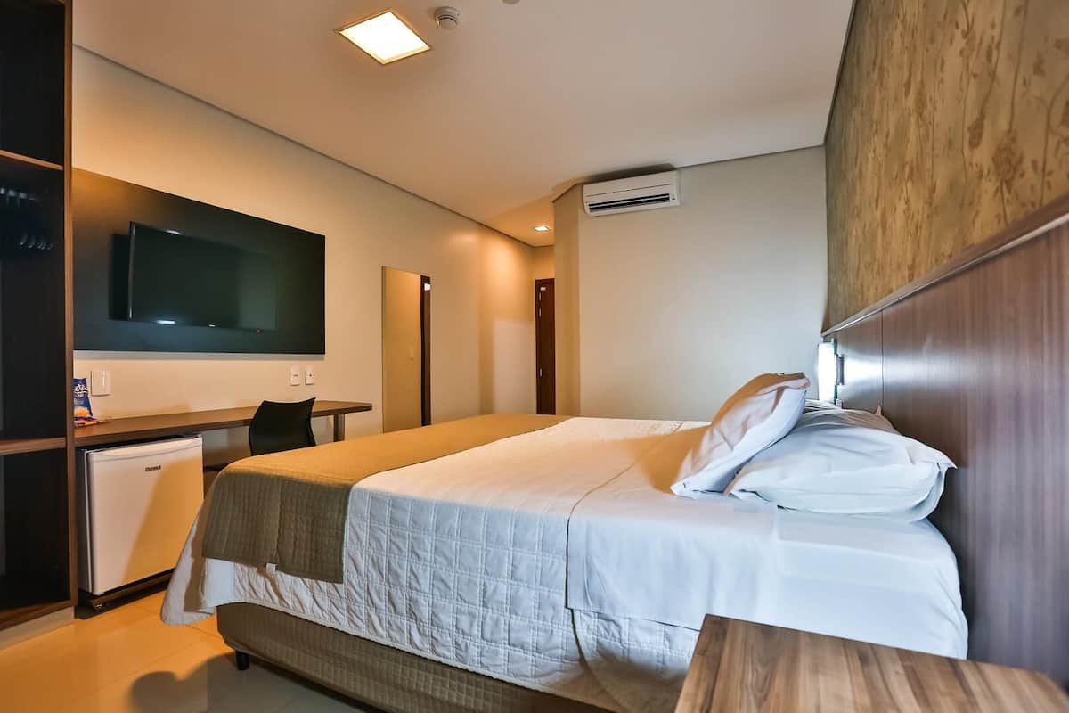 A hotel room with a bed, a desk with a chair, a television, and an air conditioning unit.