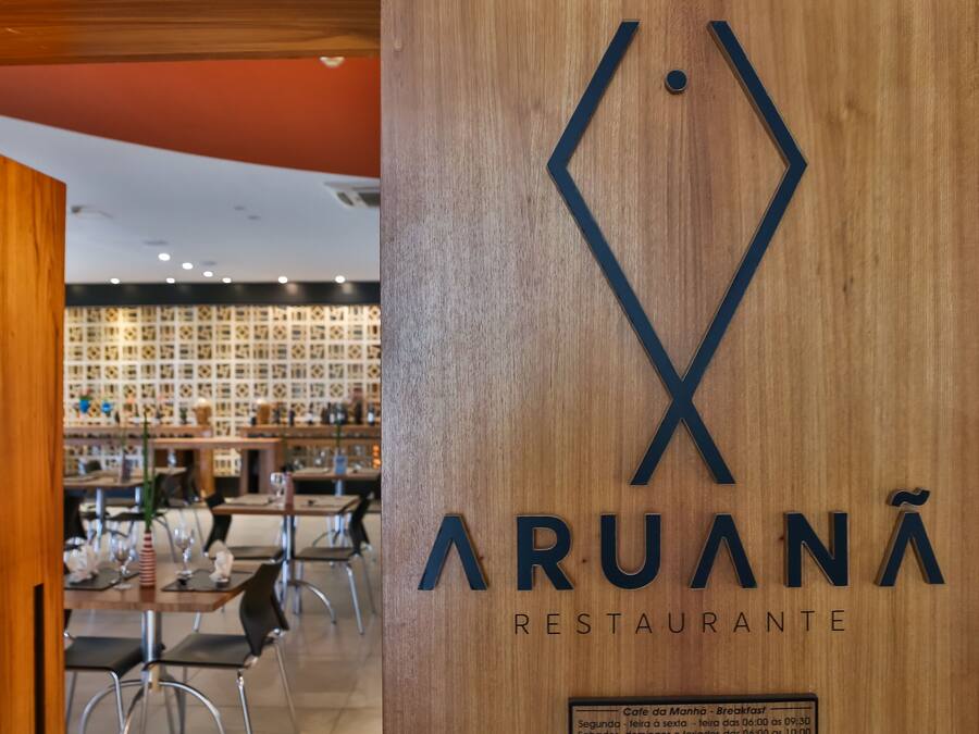 A restaurant interior with a wooden wall displaying the name 'ARUANÃ RESTAURANTE'. Tables are set with chairs, and a menu board is visible in the background.
