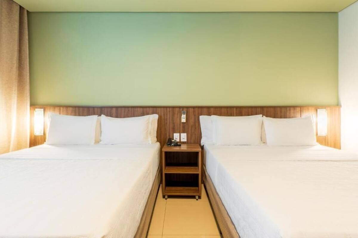 A hotel room with two single beds, wooden headboards, bedside tables with lamps, and a green wall.