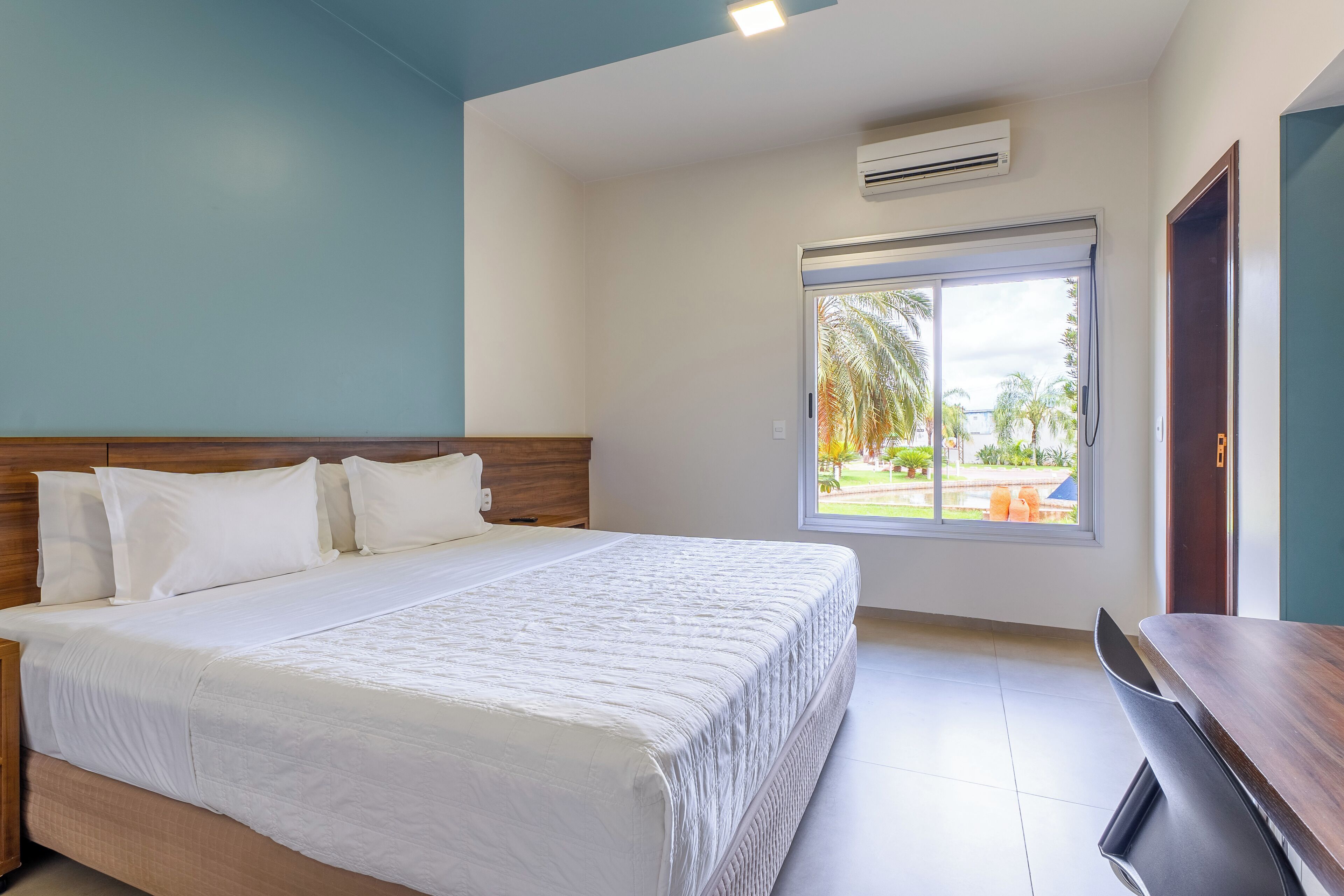 A hotel room with a bed, a desk, and a window with a view of palm trees.