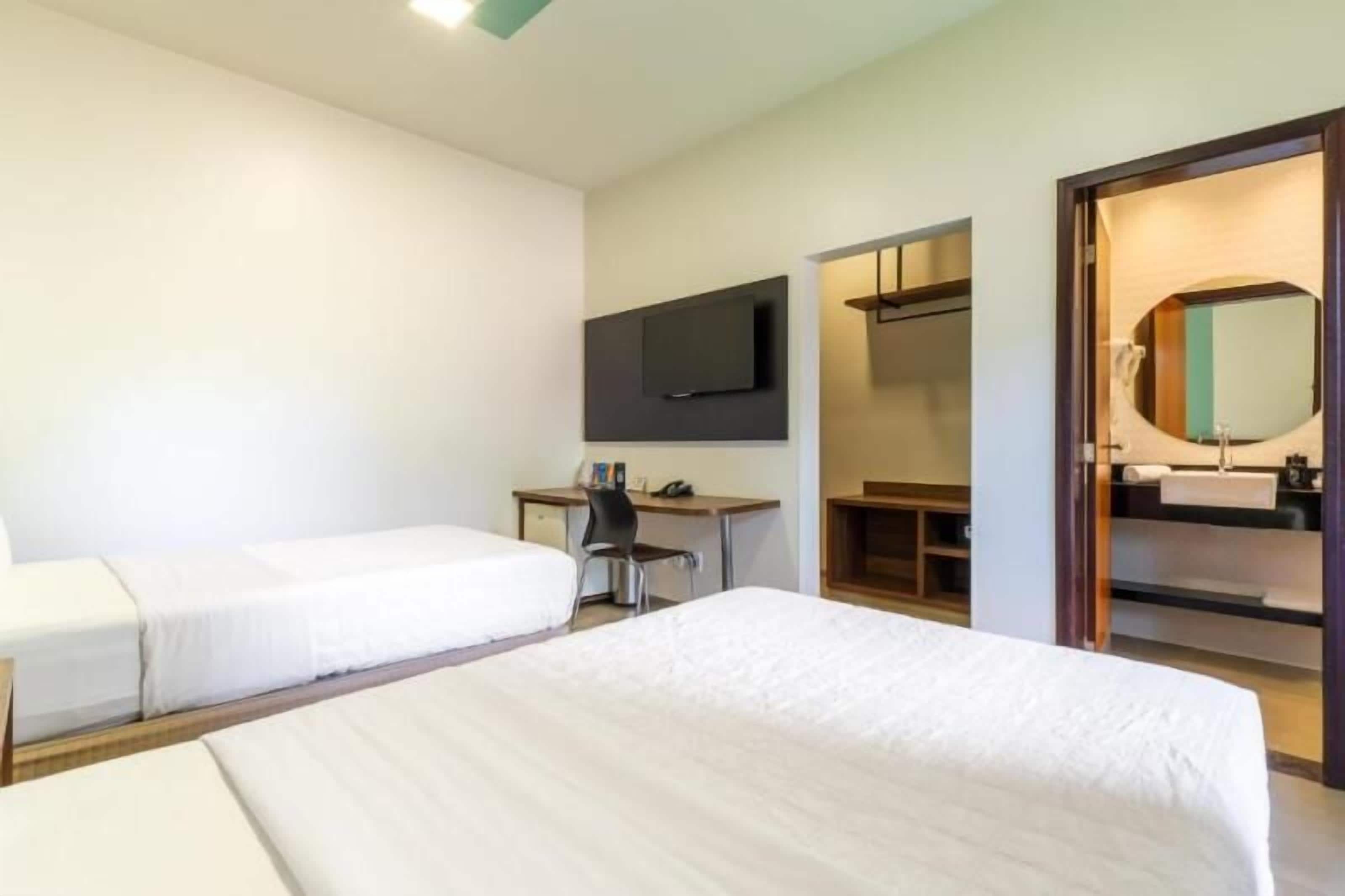 Habitación de hotel con dos camas individuales, un escritorio con silla, un televisor de pantalla plana montado en la pared, un espejo y un armario de madera.