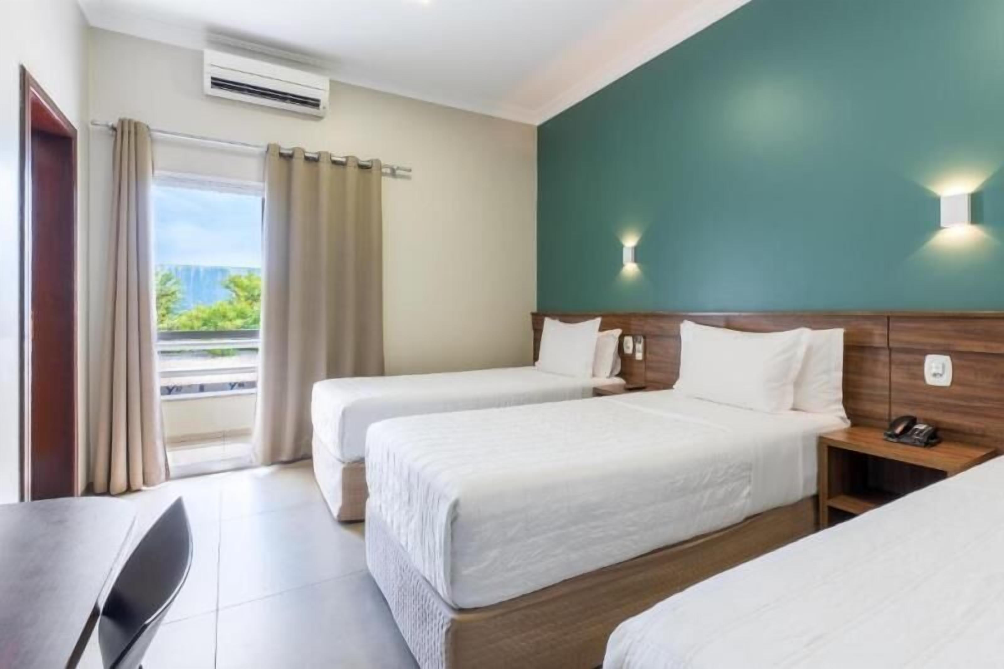 A hotel room with two beds, a desk, and a view of greenery outside.