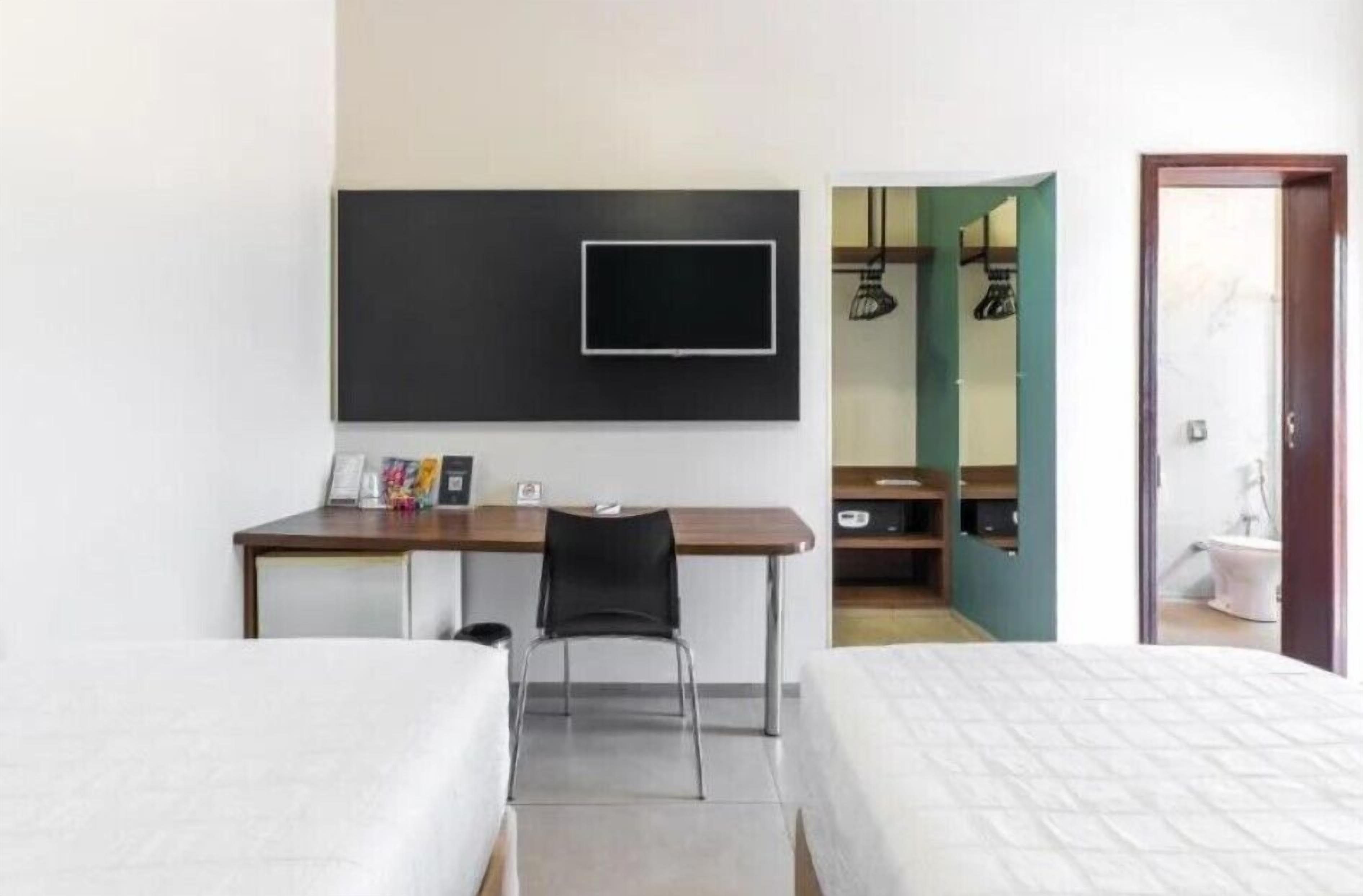 A hotel room with a bed, a desk with a chair, a TV mounted on the wall, and a bathroom visible through an open door.