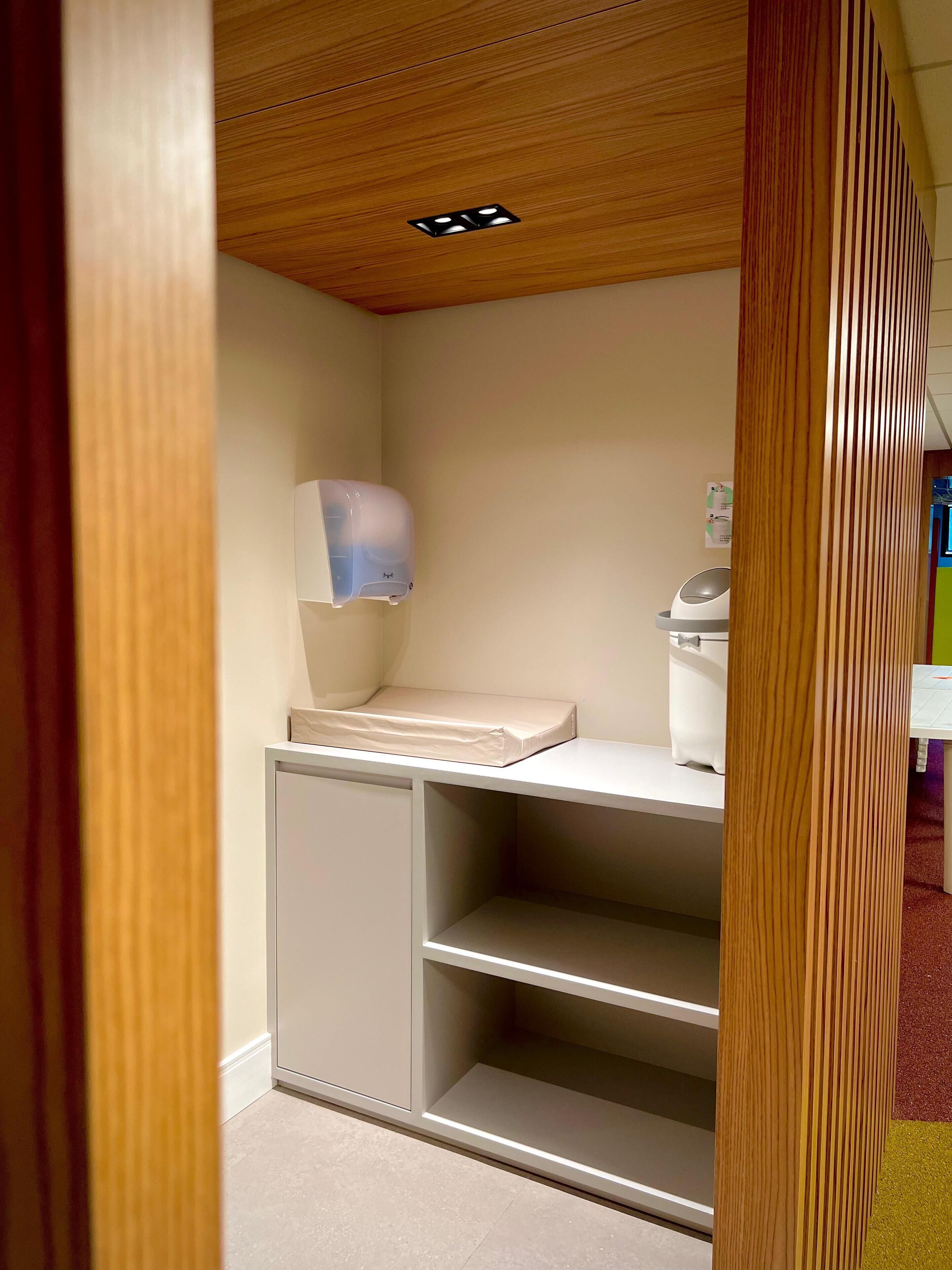 A small, compact room with a wooden ceiling, a white shelf unit, a baby changing station, and a small white cabinet.