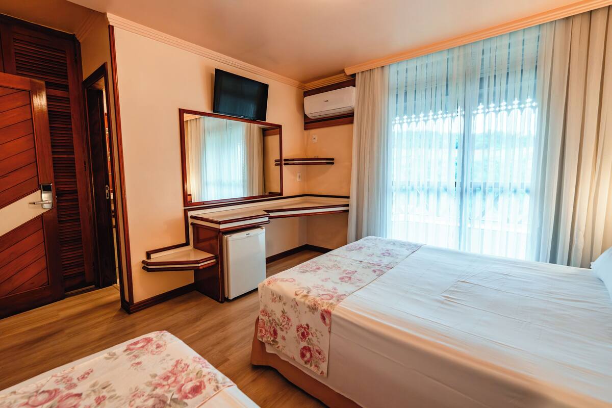 A hotel room with a bed, a desk, a mirror, a TV, and an air conditioning unit.
