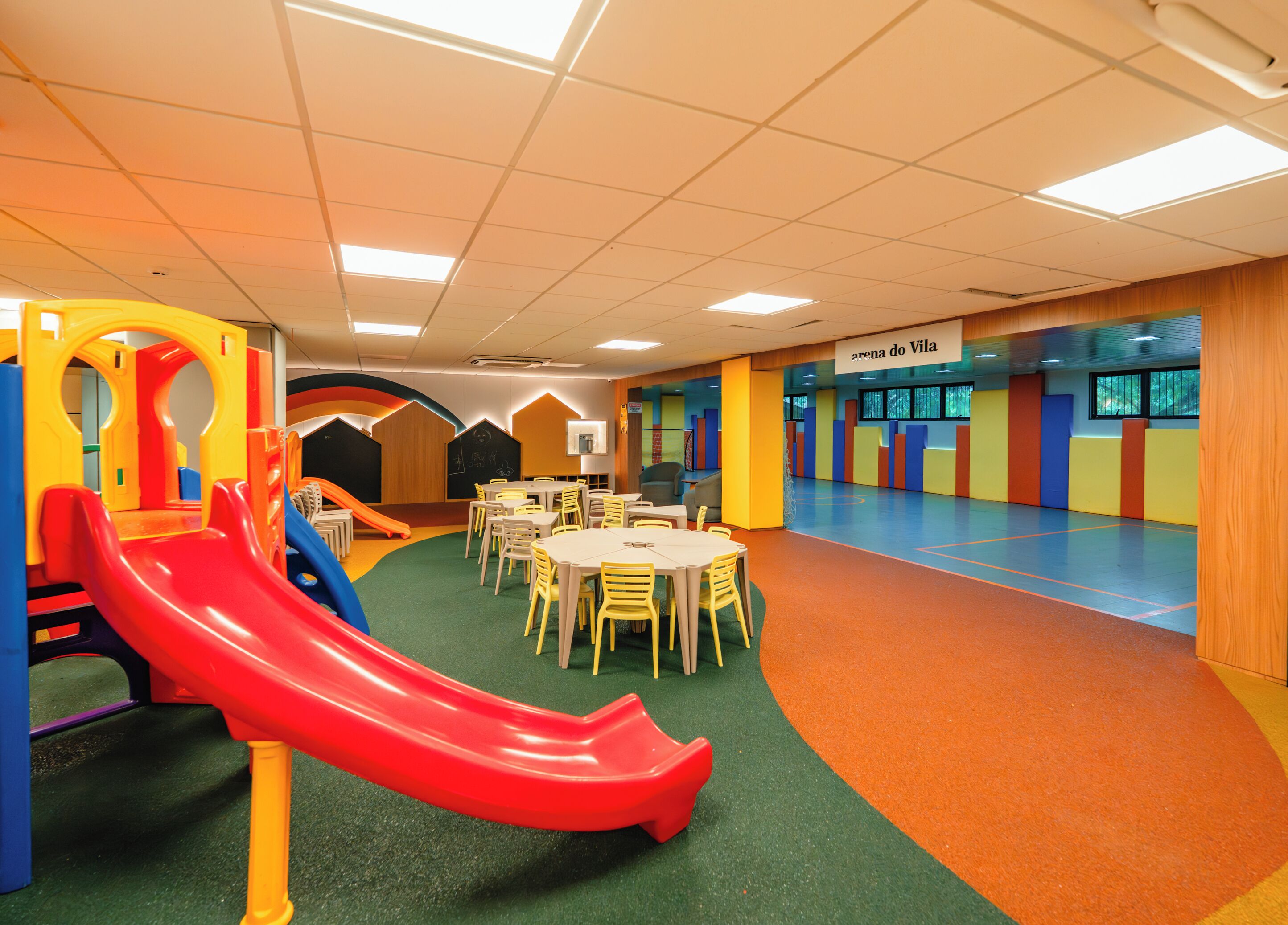 Un área de juegos para niños con un tobogán rojo, mesas y sillas, y un cartel que dice "arena do Vila".