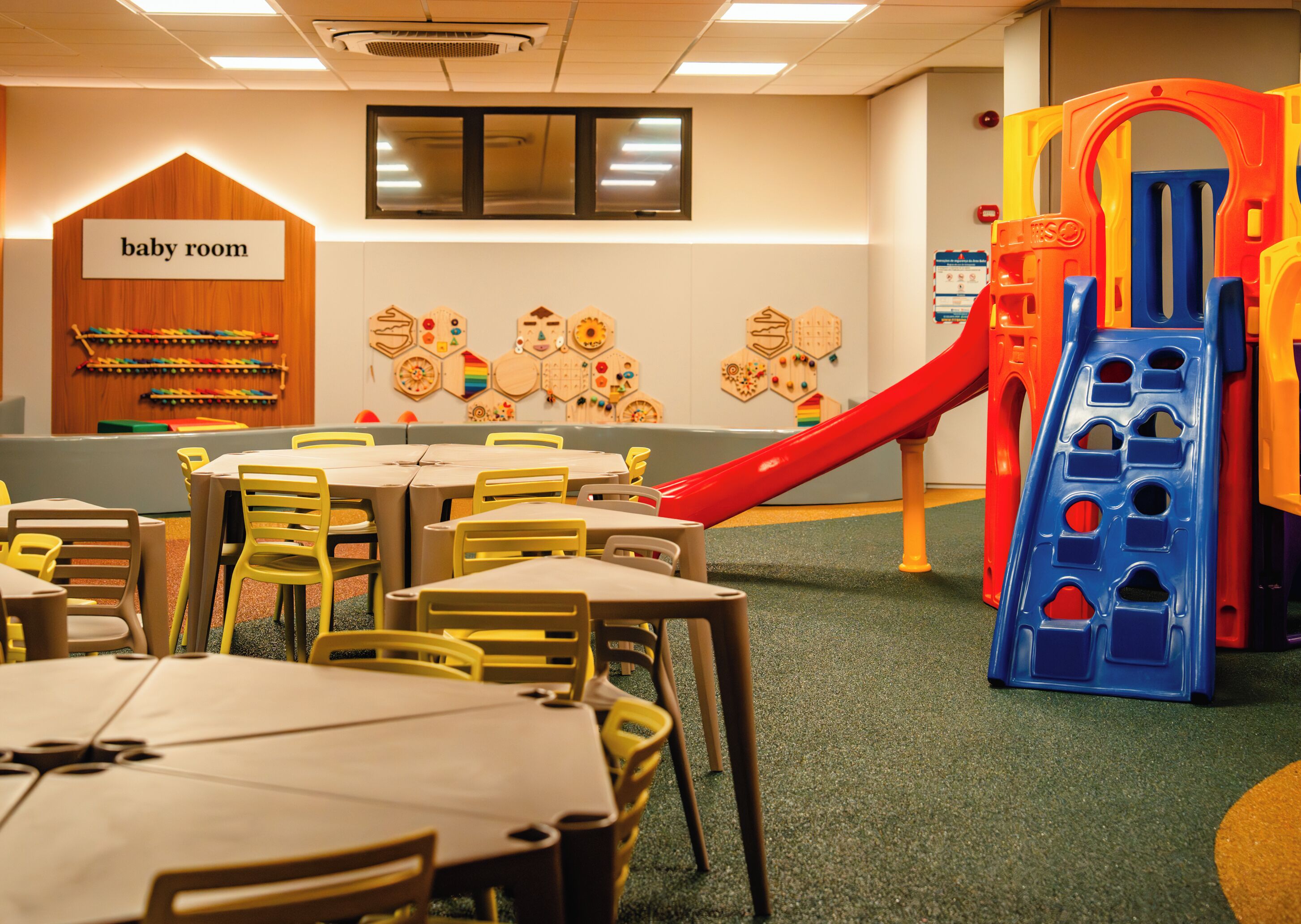 Une salle pour bébés avec un espace de jeux, des tables, des chaises et un mur décoré d’éléments éducatifs.