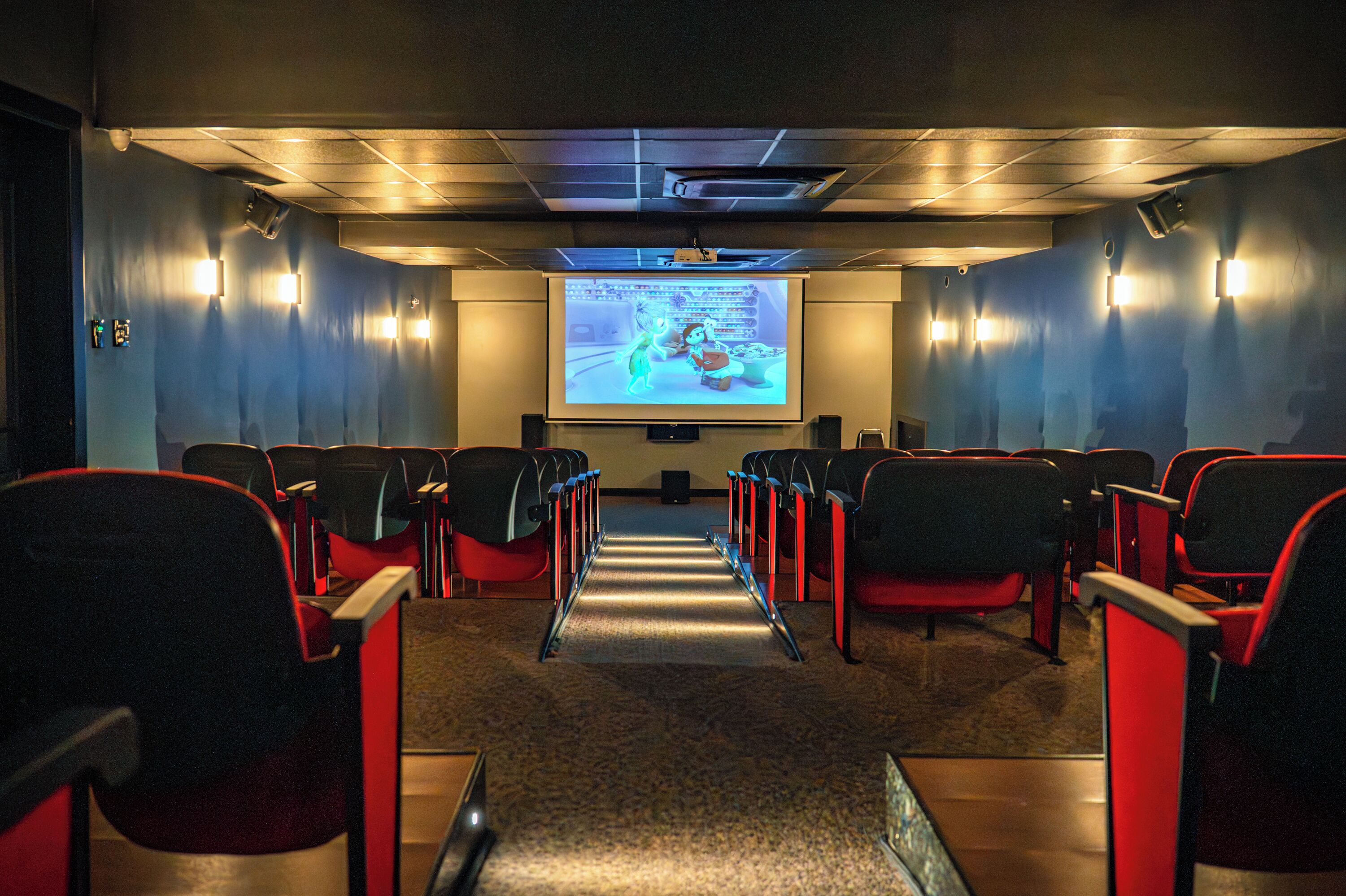 Une salle de cinéma avec des rangées de sièges rouges et noirs, un grand écran à l’avant et un sol recouvert de moquette.