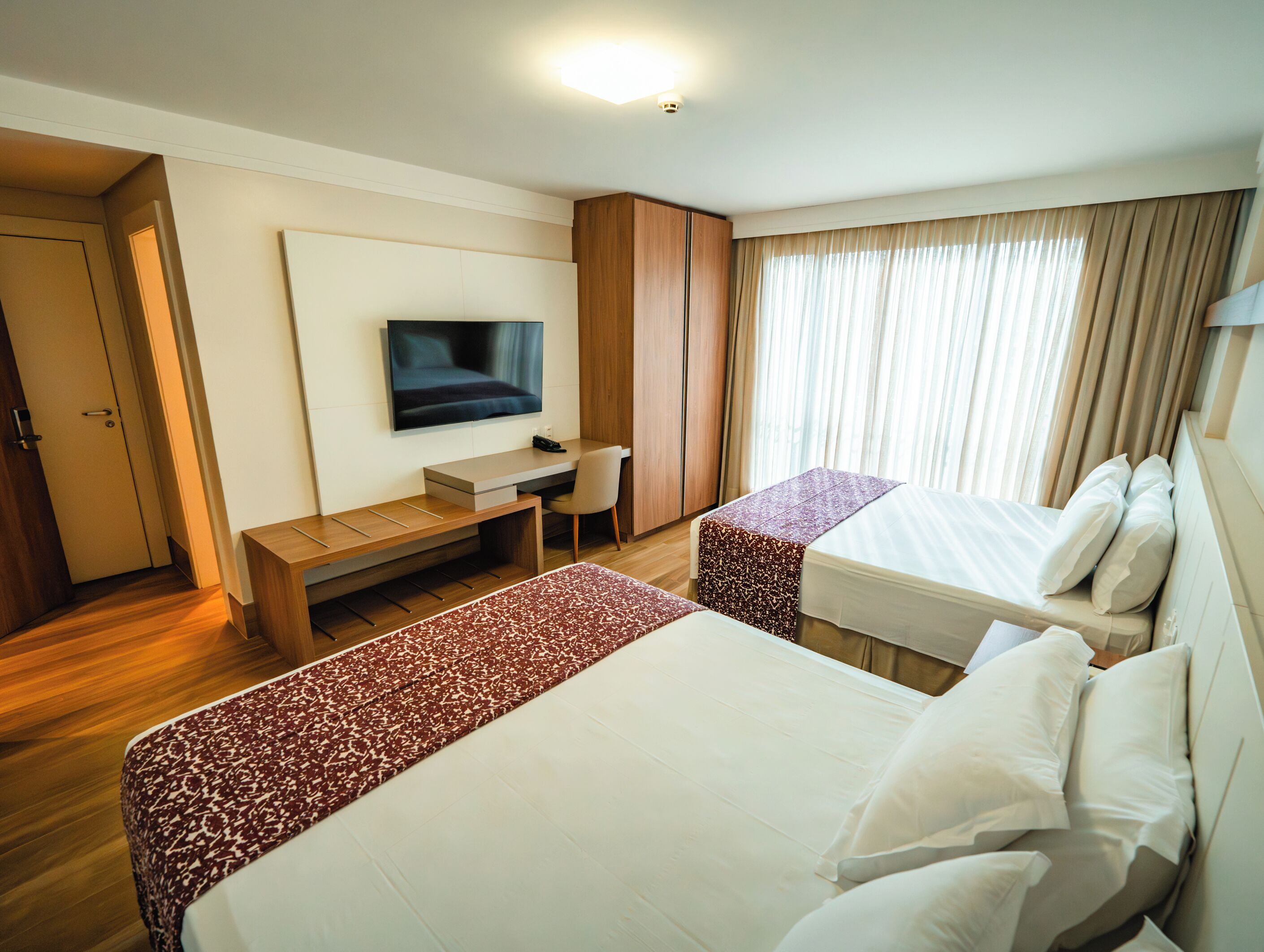 A hotel room with two beds, a desk, a television, and a window with curtains.