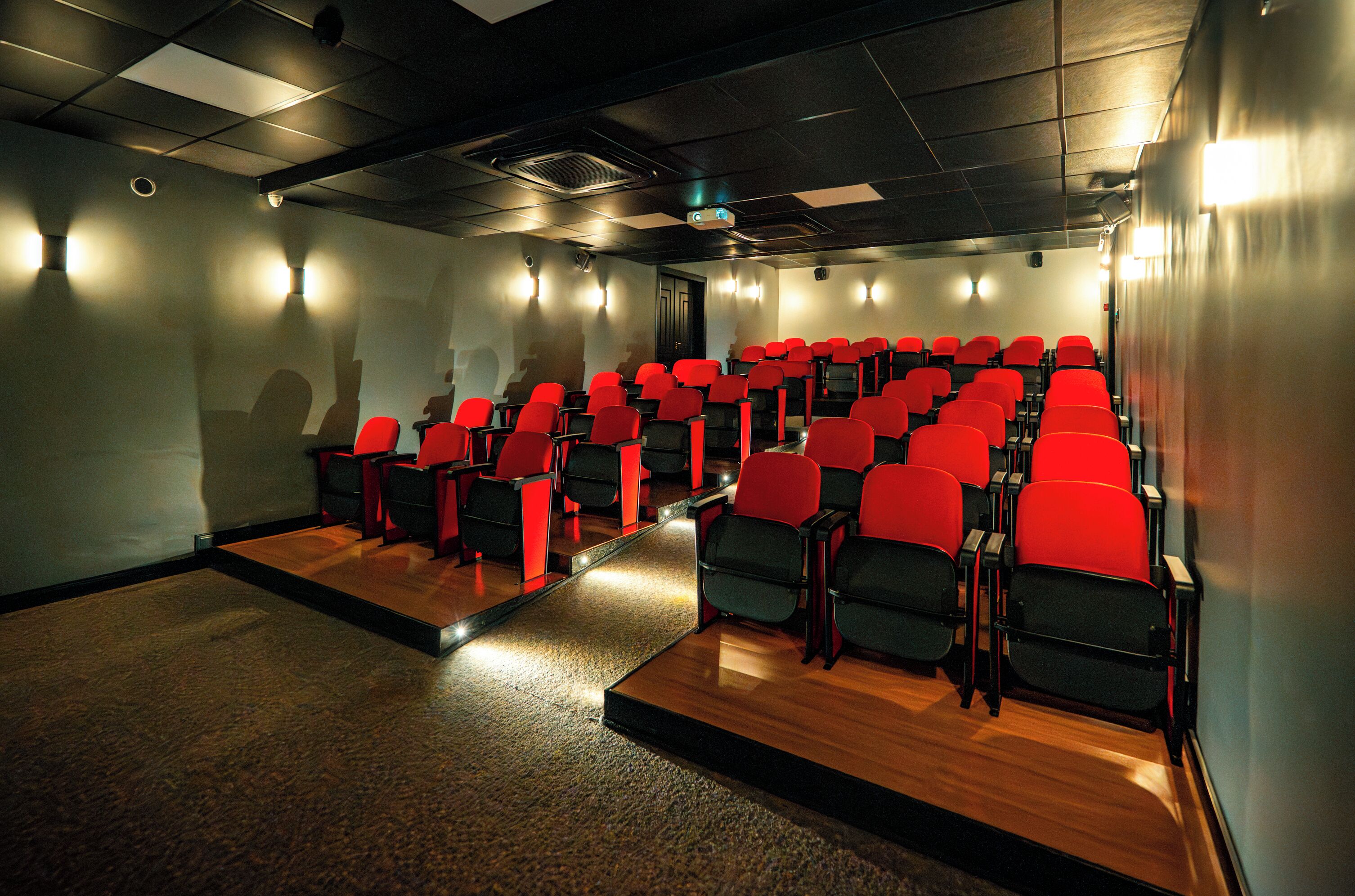 Um teatro com fileiras de assentos vermelhos e pretos, piso de madeira e uma tela de projeção.