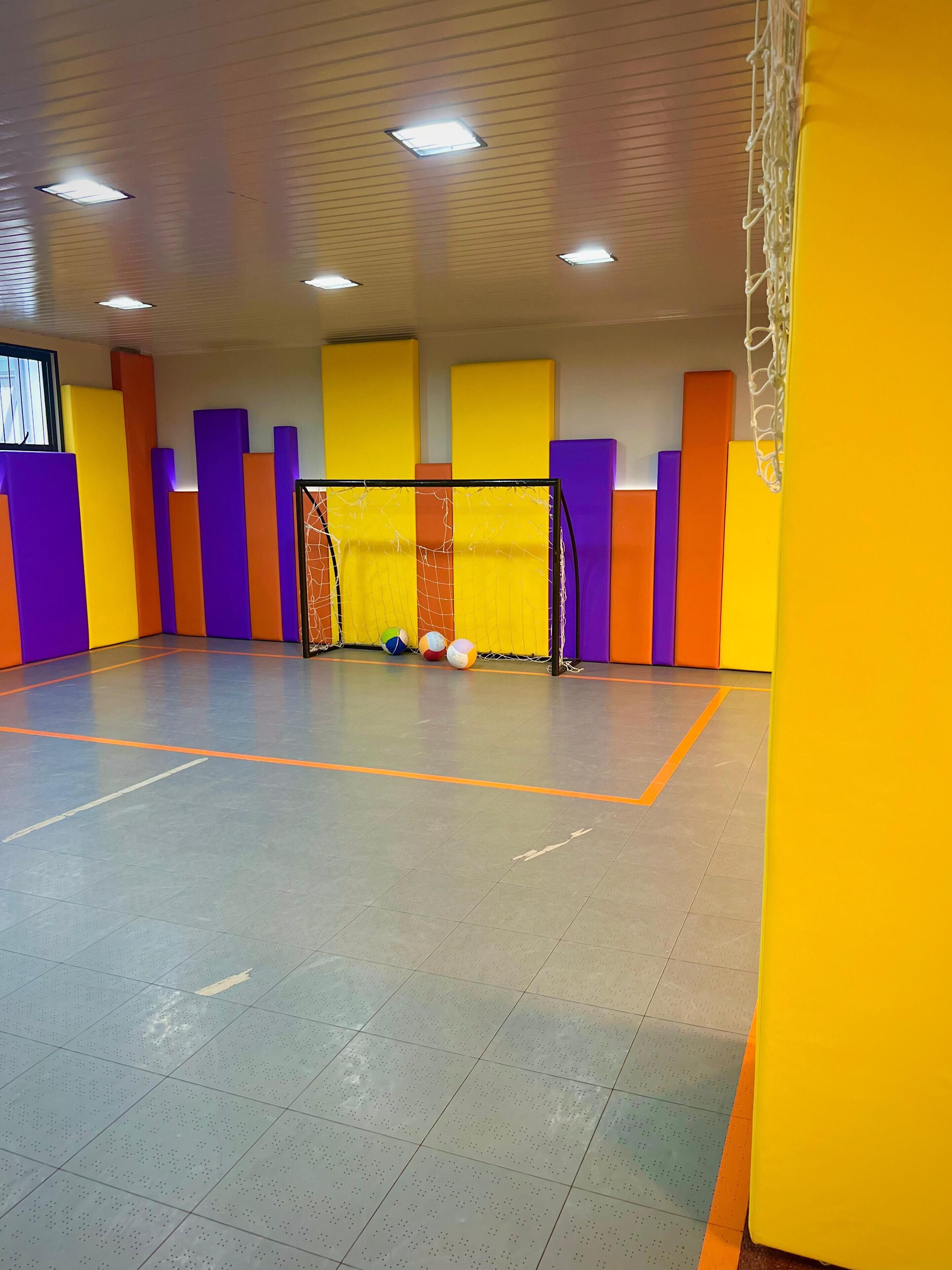 An indoor sports facility with a basketball court, a netted goal, and colorful padded walls.