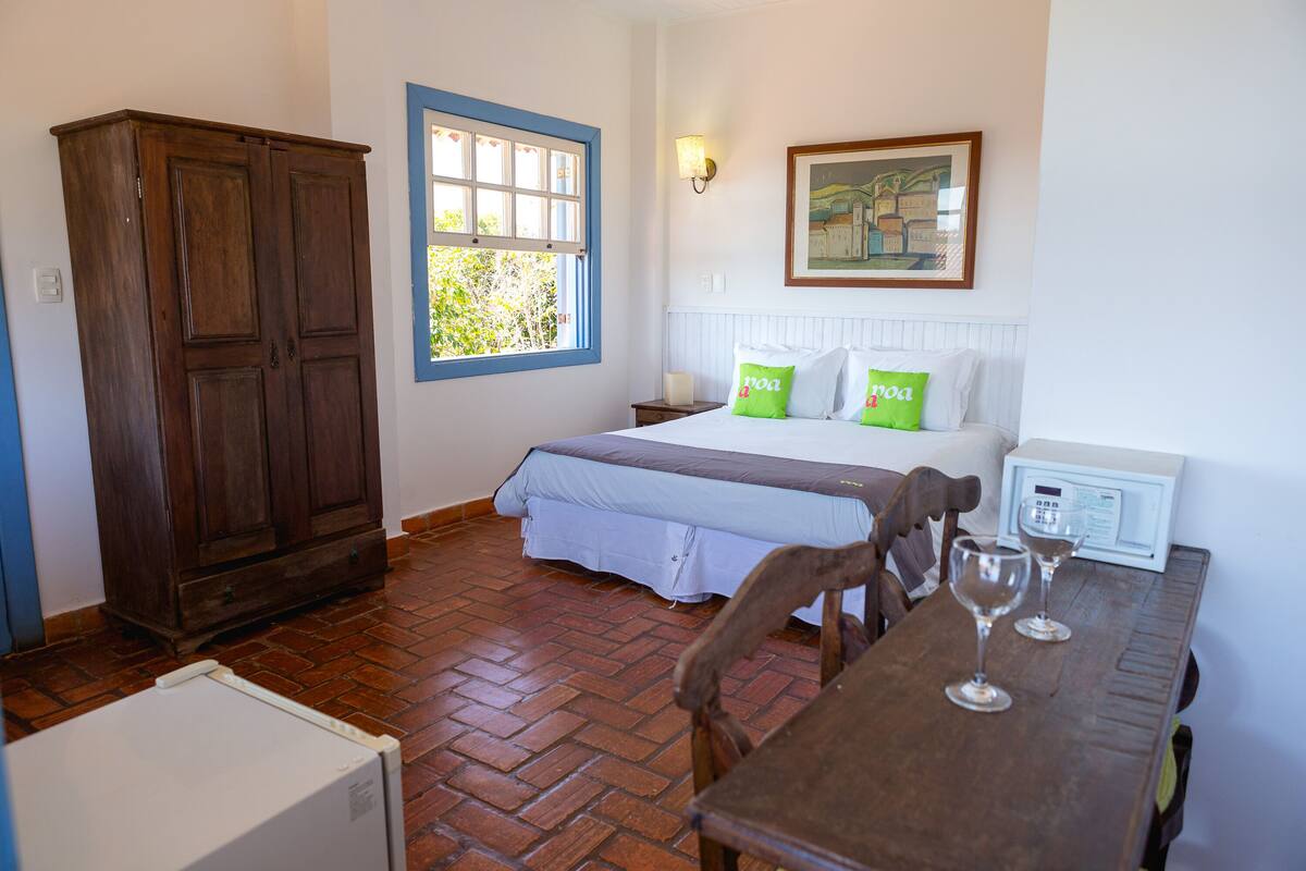 A bedroom with a bed, two pillows, a nightstand, a lamp, a wooden wardrobe, a window, and a table with two wine glasses.