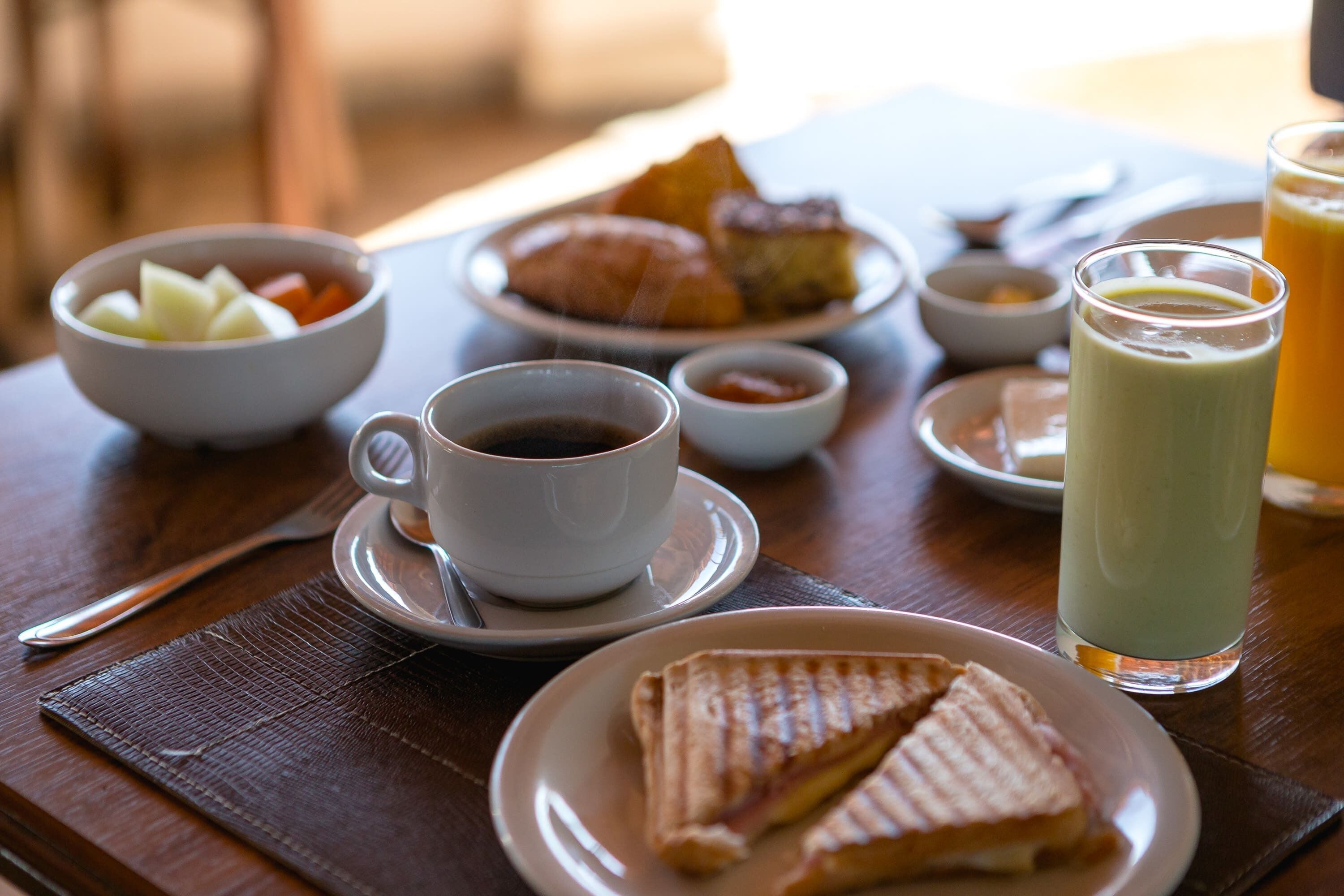 Uma mesa de café da manhã com xícara de café, copo de suco de laranja, prato de croissants e tigela de frutas.