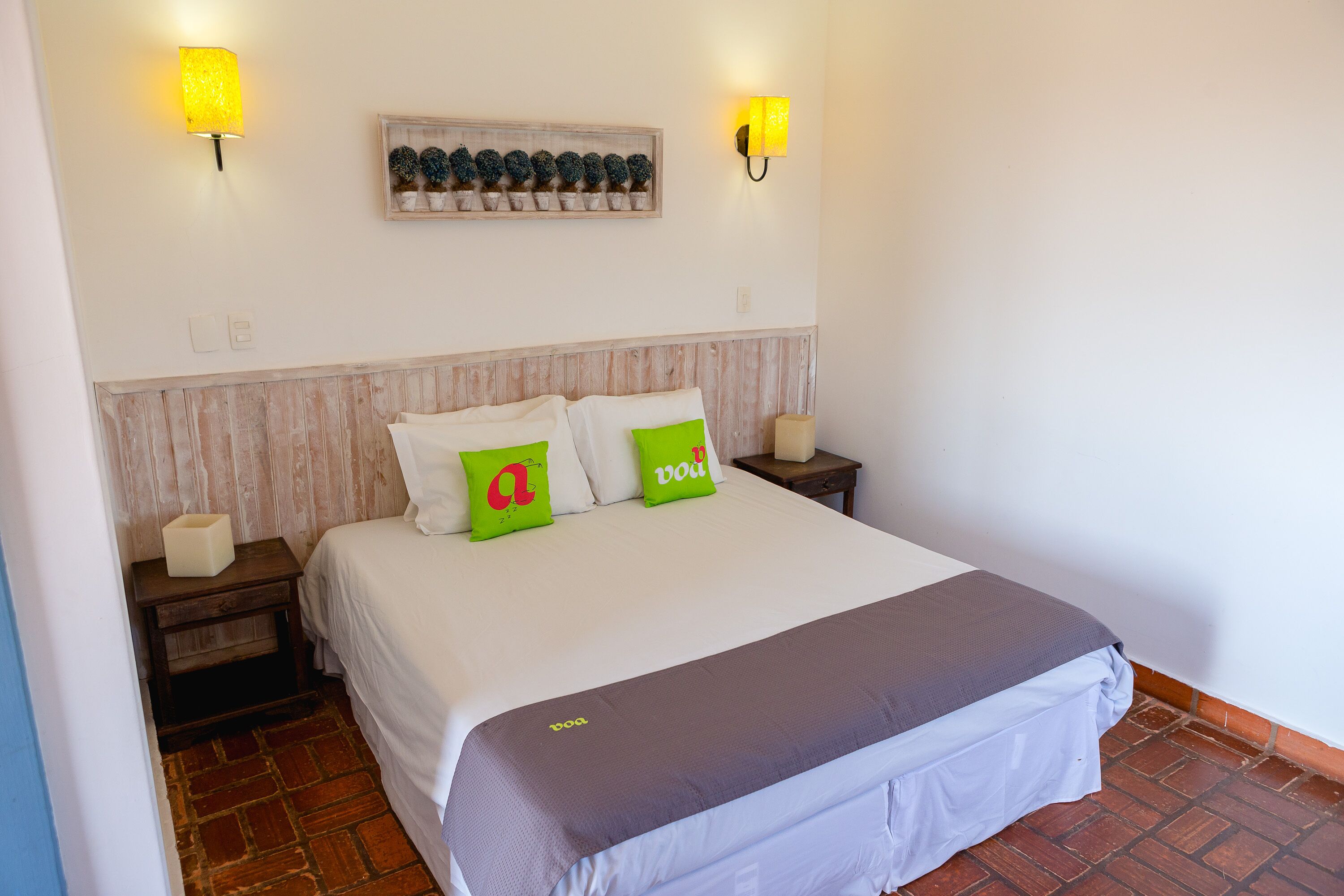 A neatly made bed with white bedding and two green pillows, a wooden headboard, a small bedside table with a lamp, and a wall-mounted shelf with decorative items.