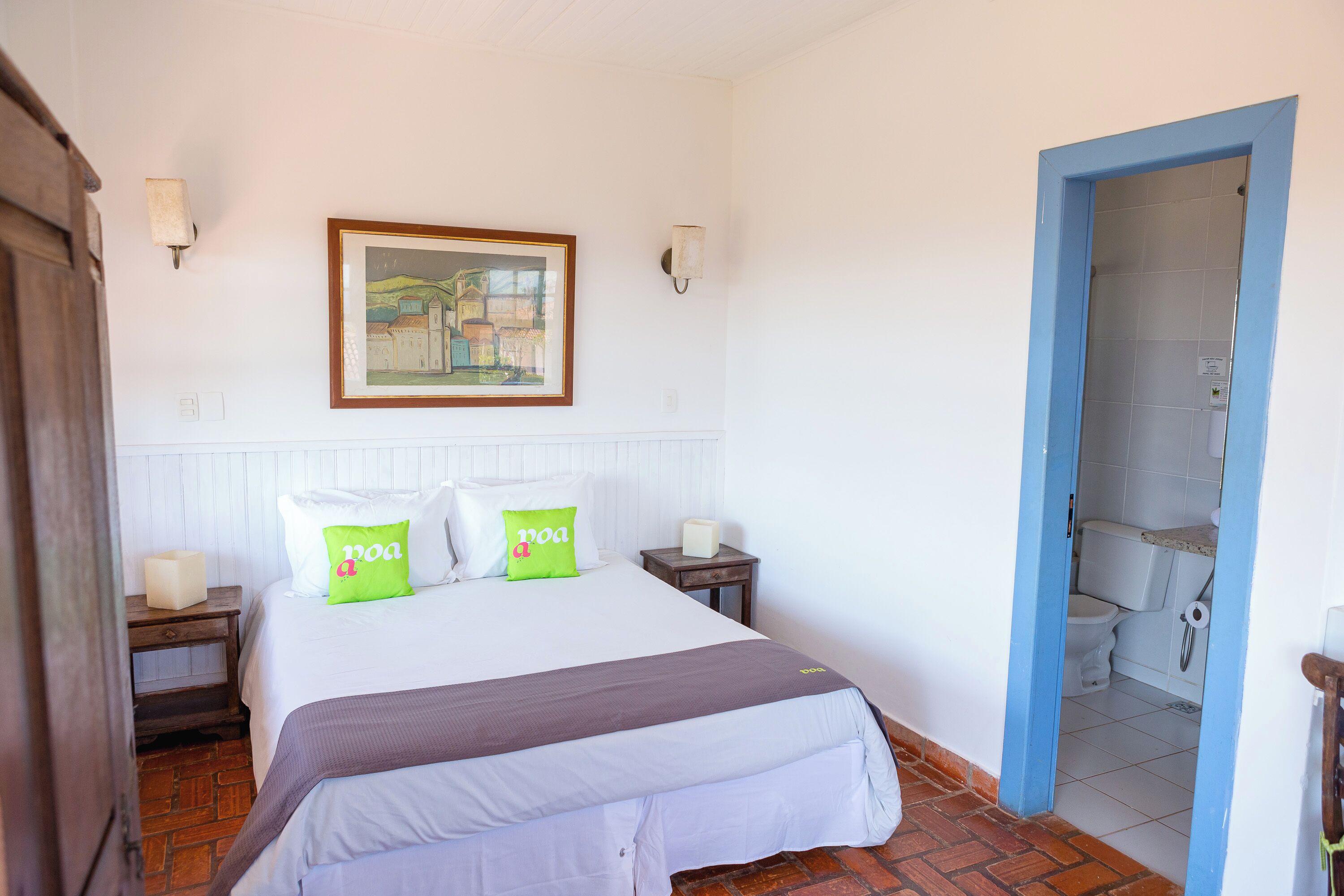 Un dormitorio con cama, dos mesitas de noche, un cuadro enmarcado en la pared y la puerta del baño visible a través de un vano abierto.
