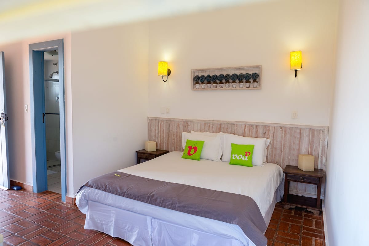 A neatly made bed with two pillows, a bedside table with a lamp, and a wall-mounted shelf with decorative items.