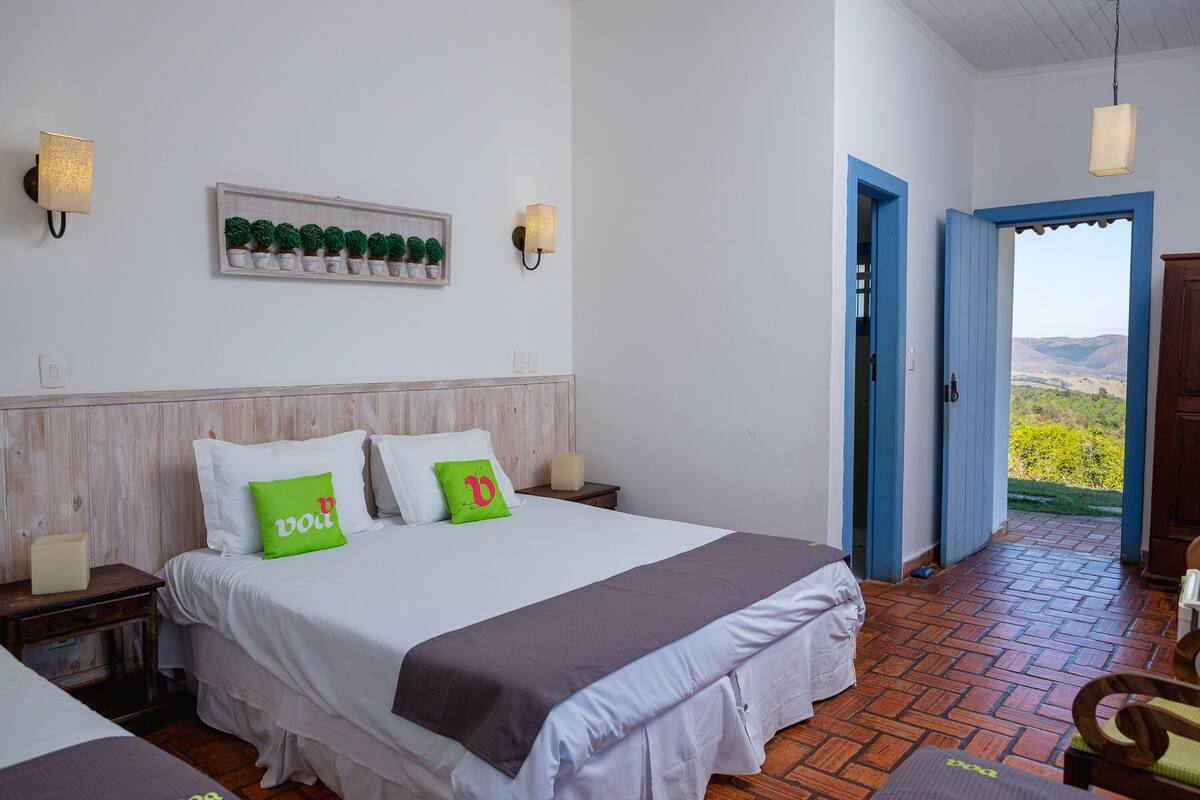 A bedroom with a bed, bedside tables, a wall-mounted shelf with potted plants, and a view through an open door.