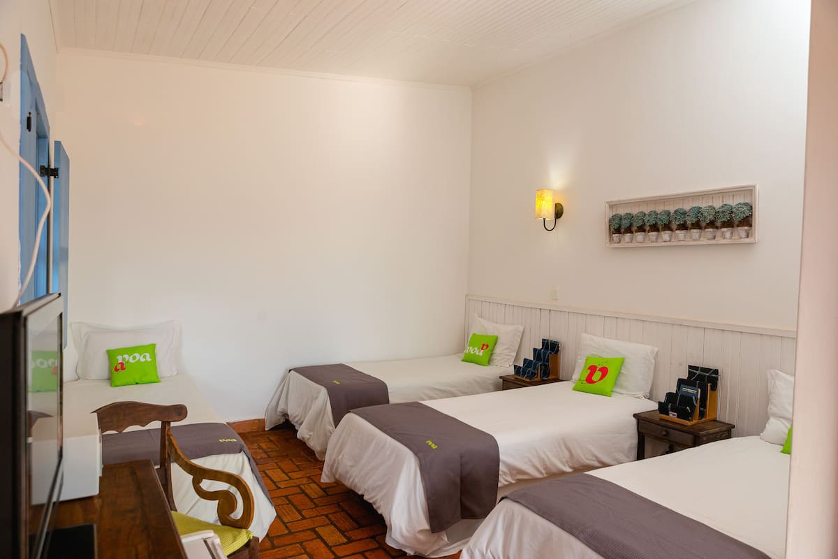 A hotel room with three beds, a desk, a chair, a nightstand, a lamp, and a shelf with decorative items.