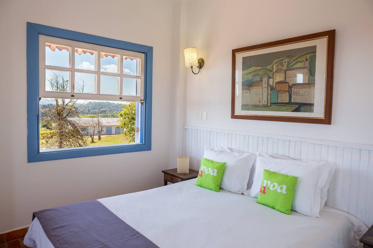 A bedroom with a bed, pillows, a nightstand, a lamp, a window with a view, and a framed picture on the wall.