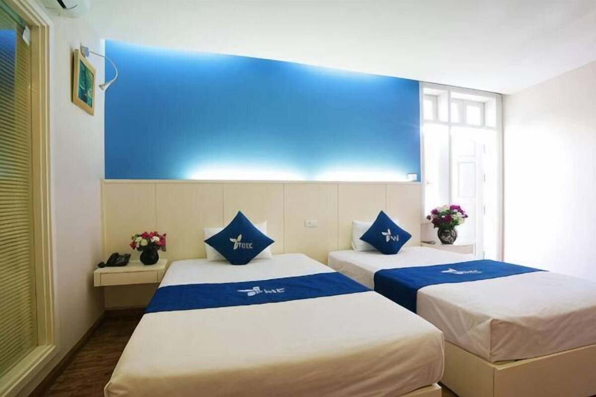 A hotel room with two beds, a bedside table with a phone, a vase with flowers, and a large blue wall.