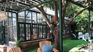 A covered outdoor seating area with wooden chairs and tables, surrounded by greenery and a glass enclosure. - Ping Hanoi Hotel (Hanoi)