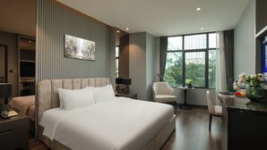 A modern hotel room with a large bed, a desk with a chair, a small table with a vase of flowers, and a large window with a city view. - Ping Hanoi Hotel (Hanoi)