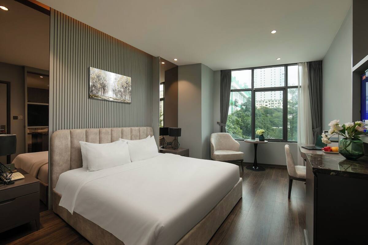 A modern hotel room with a large bed, a desk with a chair, a small table with a vase of flowers, and a large window with a city view.
