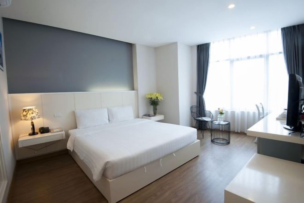 A modern hotel room with a large bed, bedside table, lamp, and a small desk area with a chair.