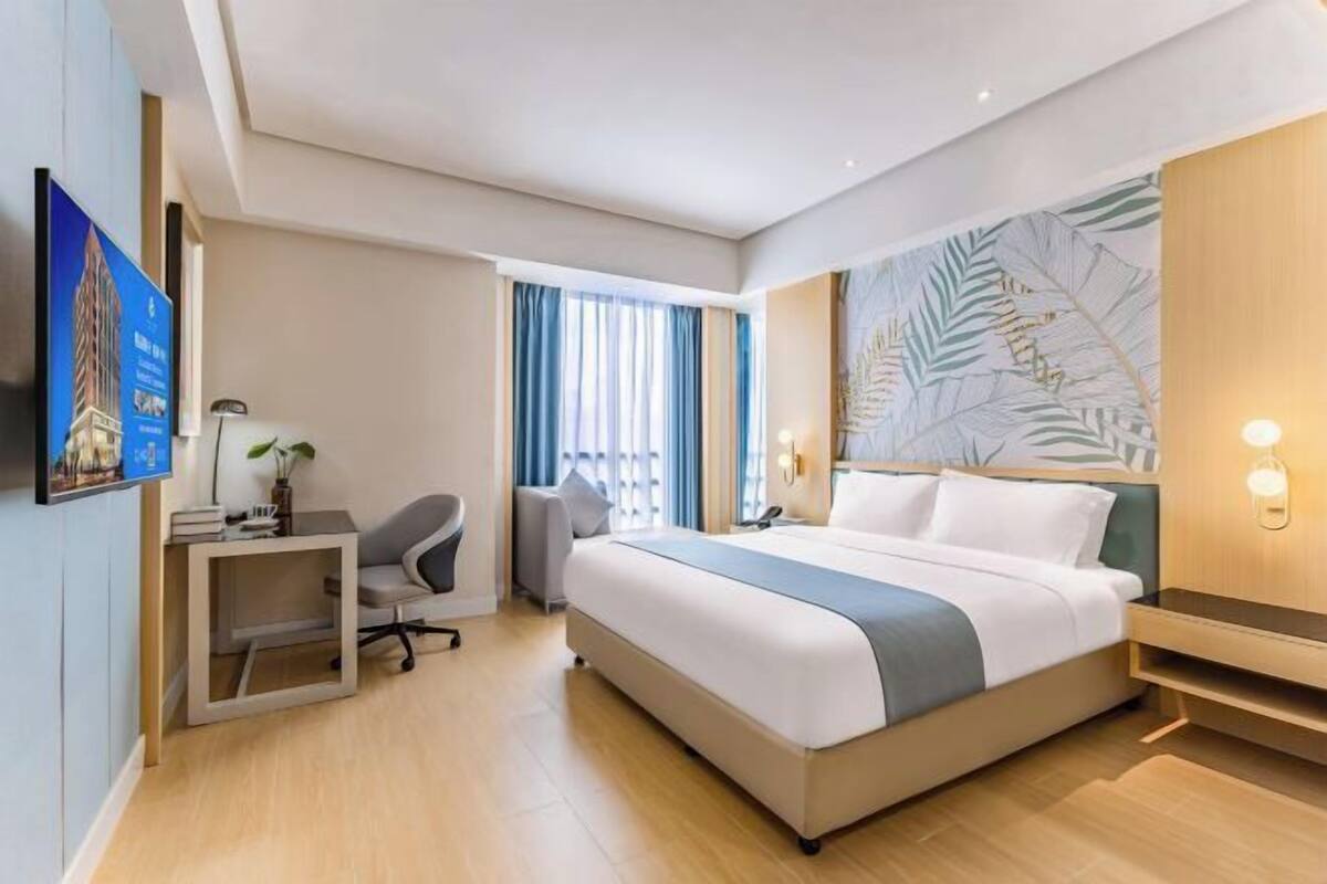 Una habitación de hotel moderna con una cama grande, un escritorio con silla, una mesita auxiliar pequeña, un televisor montado en la pared y una ventana con cortinas.