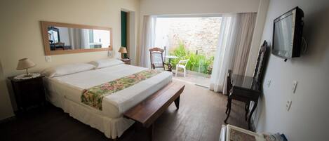 Hotelkamer met een bed, een bankje, een bureau met een televisie, een stoel en uitzicht op de tuin door het raam.