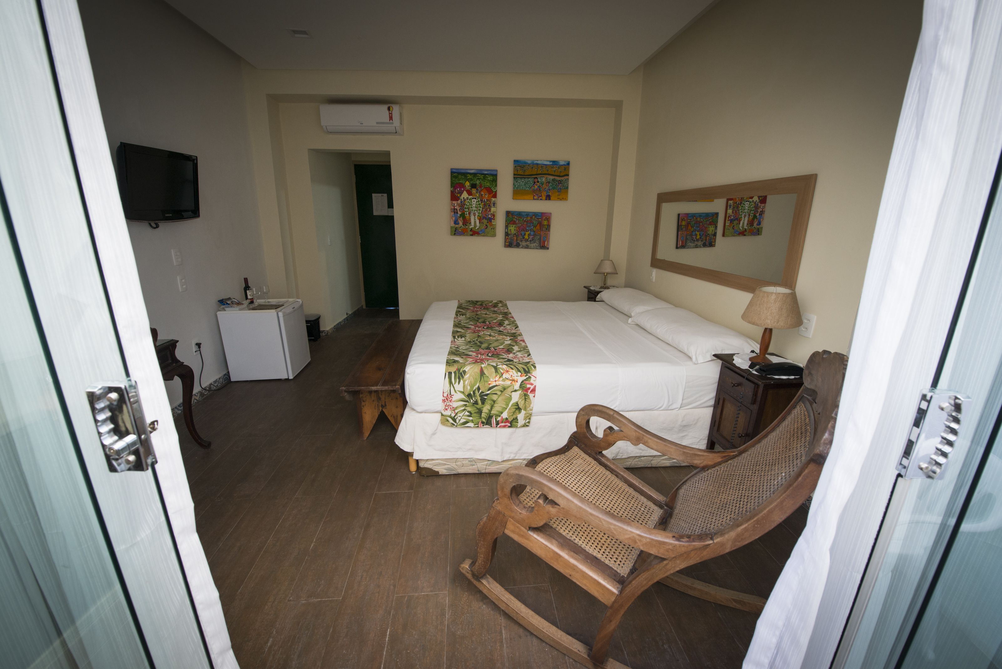 A hotel room with a bed, a wooden rocking chair, a small table, a television, and artwork on the walls.