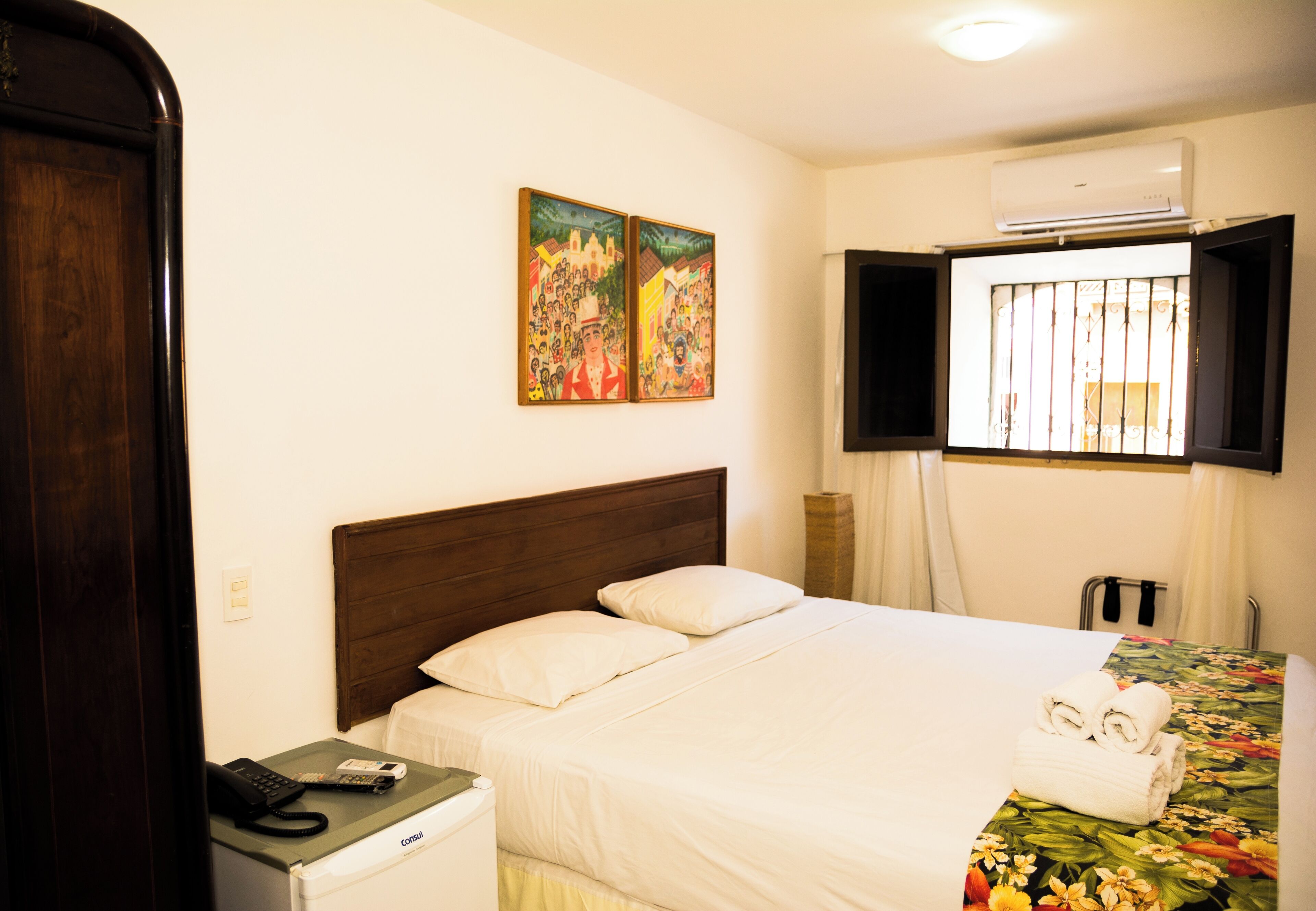 Une chambre d’hôtel avec un lit, une table de chevet avec un téléphone et une télécommande, une fenêtre avec des rideaux et deux tableaux au mur.