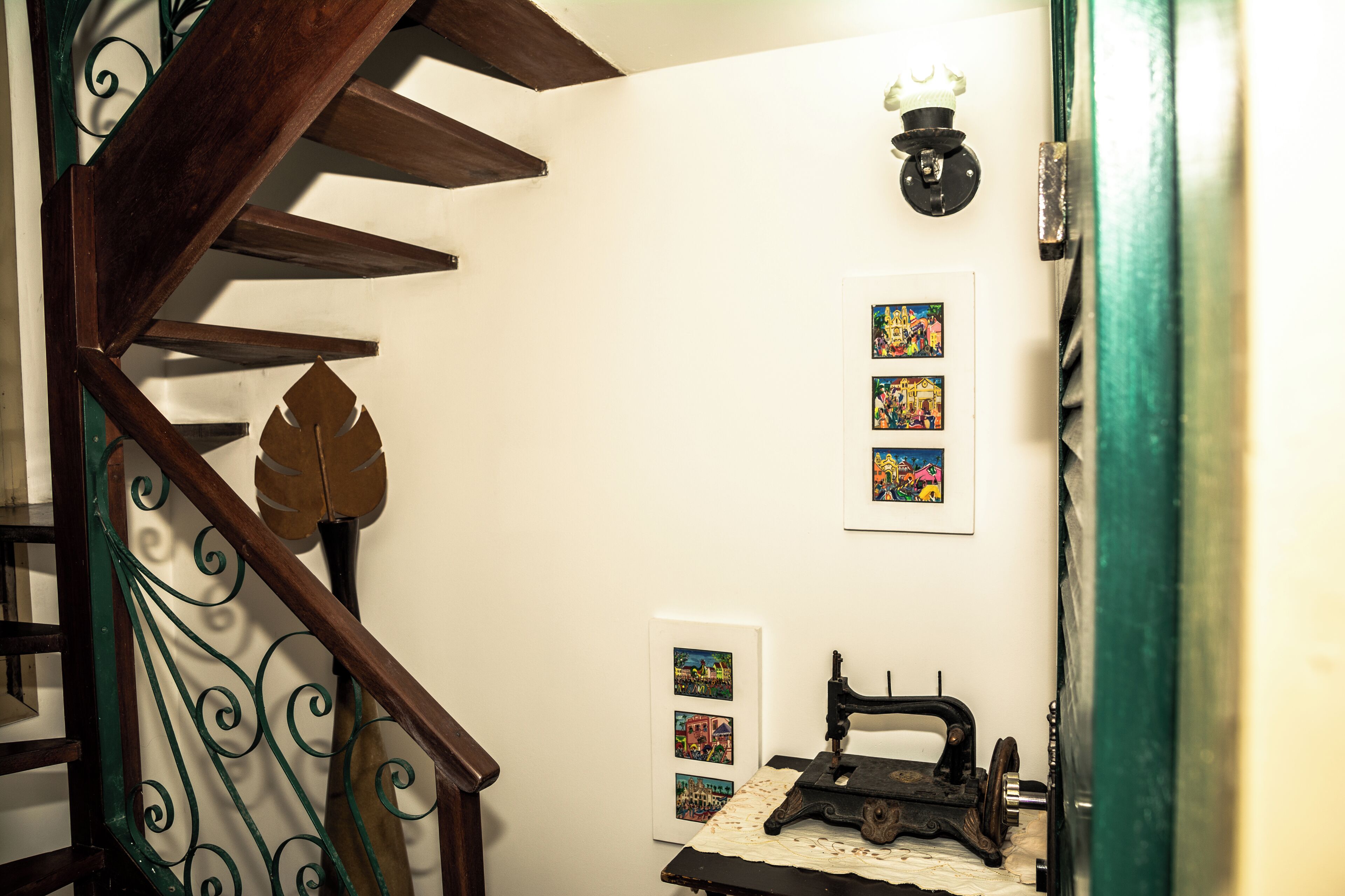 Una escalera con barandilla de madera y un adorno con forma de hoja. En la pared hay cuadros enmarcados y una máquina de coser sobre una mesa.