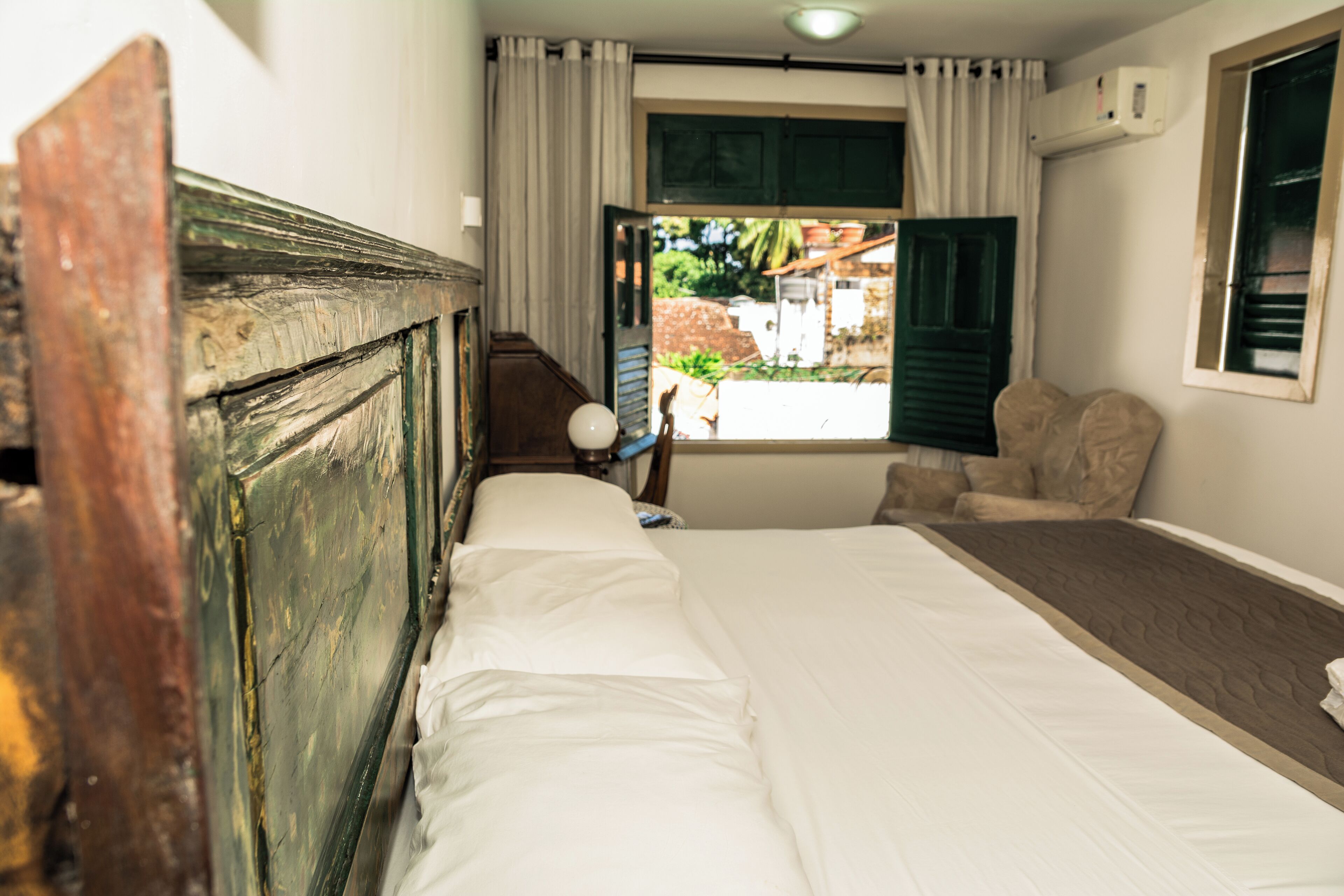 Una habitación con cama, una silla, un ventanal con cortinas y un equipo de aire acondicionado.