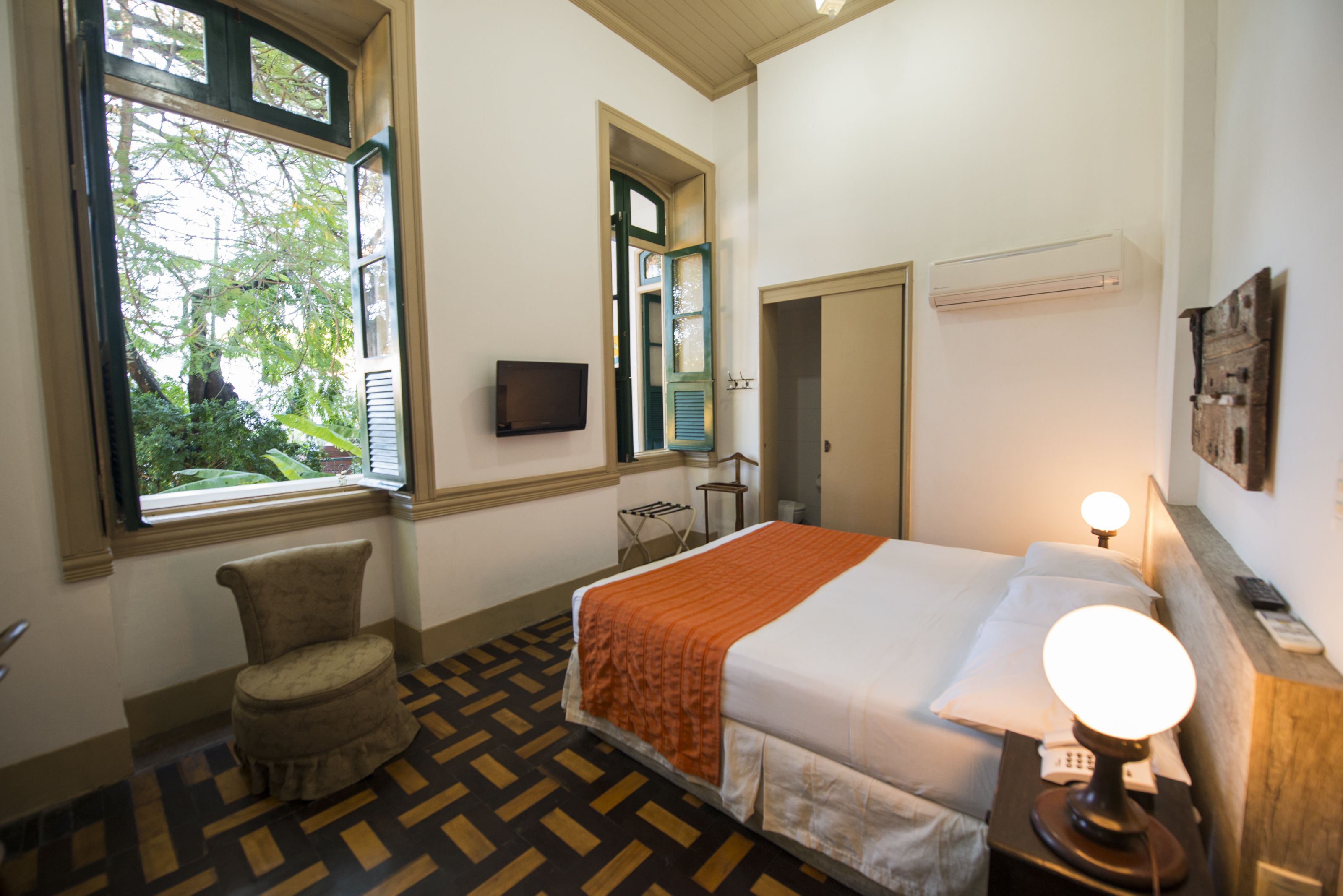 Habitación de hotel con cama, una silla, televisor, ventana y mesita de noche con lámpara.