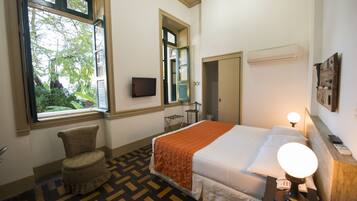 Habitación de hotel con cama, una silla, televisor, ventana y mesita de noche con lámpara.
