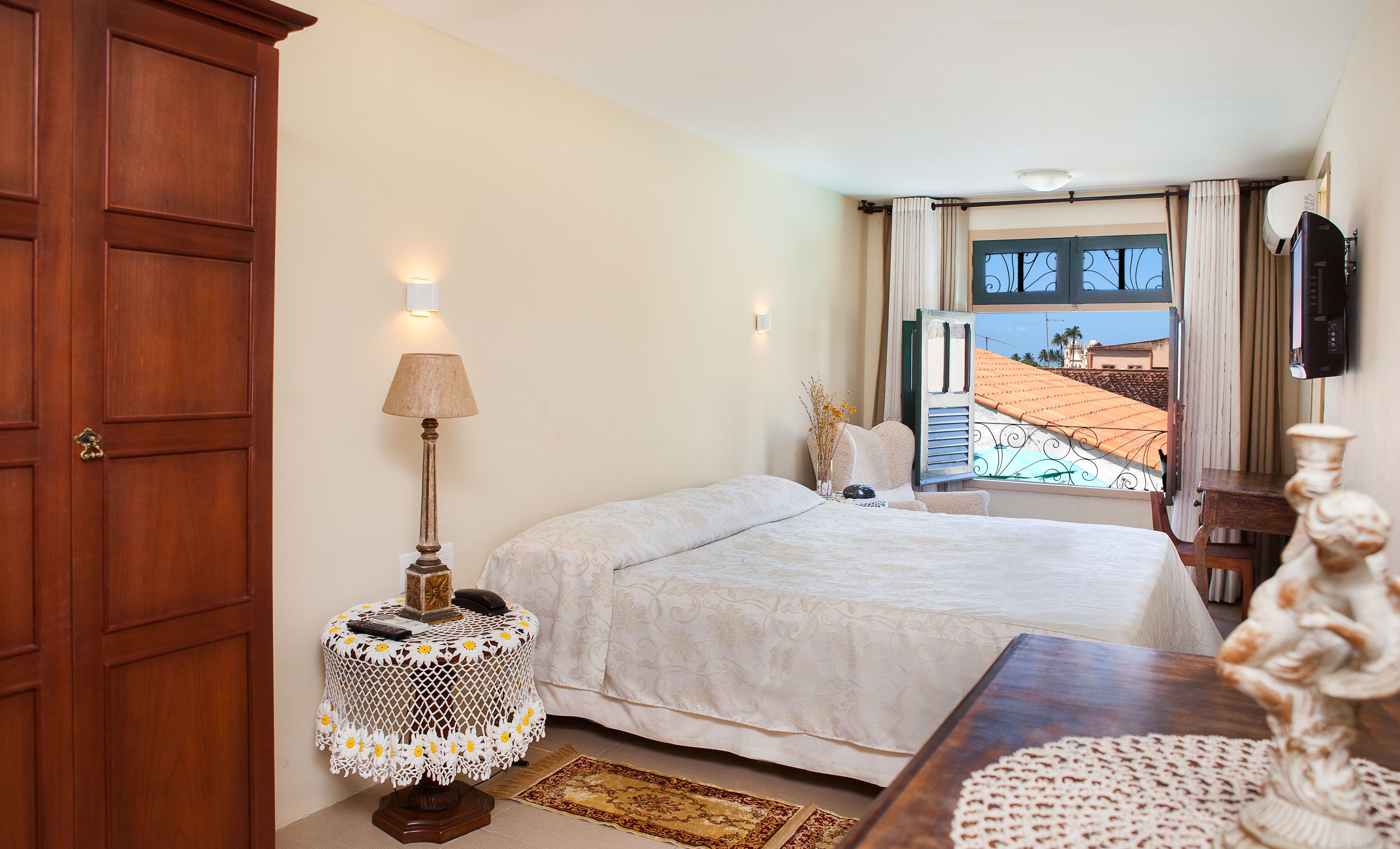 A hotel room with a bed, a bedside table with a lamp, a view of the outside, and a wooden wardrobe.