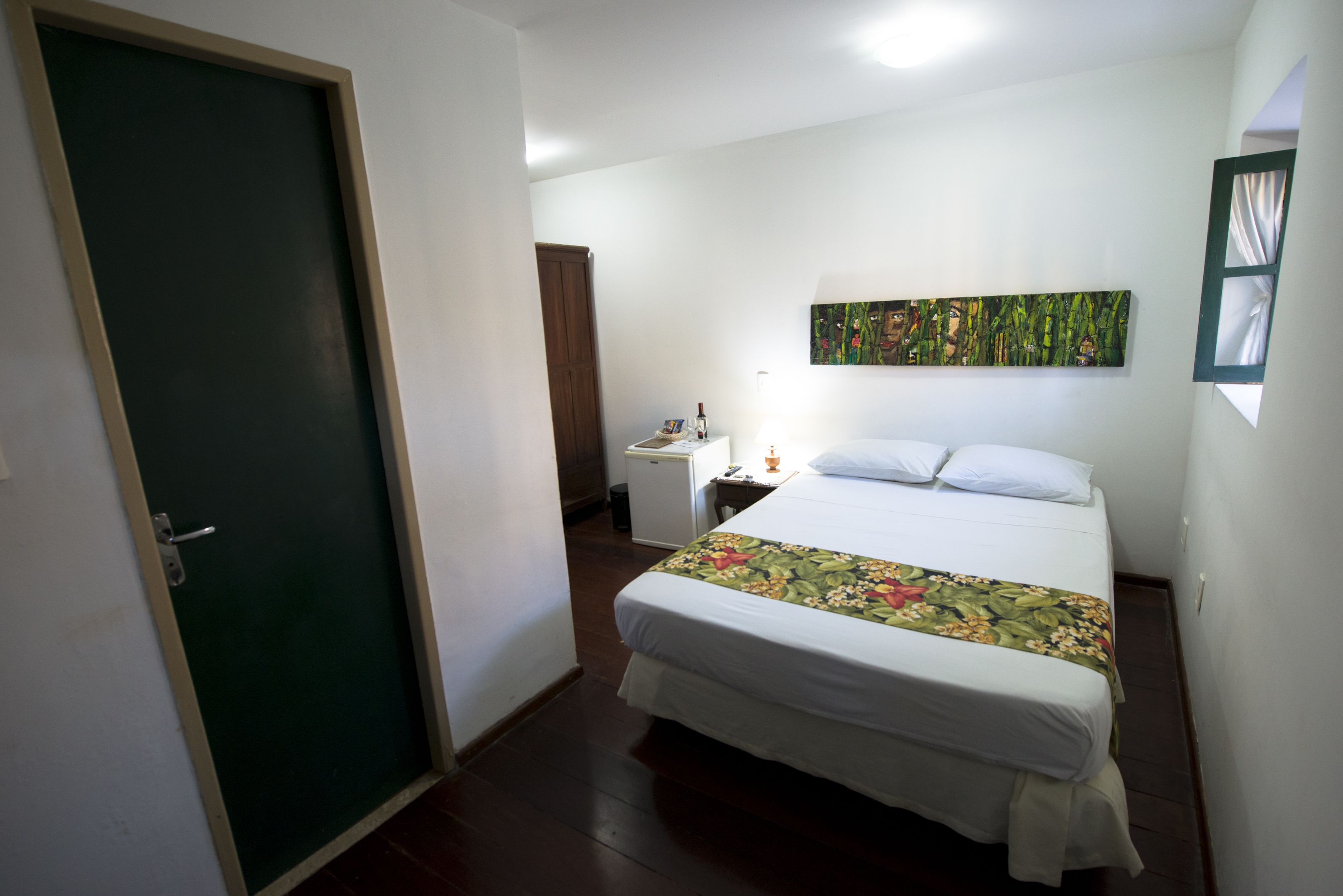 Una habitación de hotel con una cama, una mesita de noche, una pequeña mesa, una puerta y una pared con un cuadro.