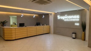 A reception desk with a digital display, a plant, and a trash can. - THE - Tropicanas Hotel e Eventos (Florianópolis)