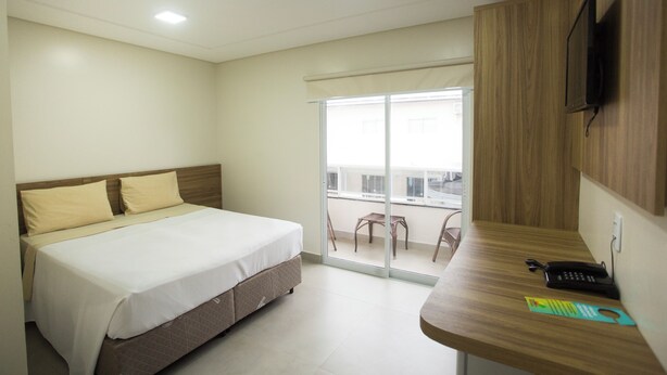 A bedroom with a bed, a desk, a chair, a TV, and a balcony with a view. - THE - Tropicanas Hotel e Eventos (Florianópolis)