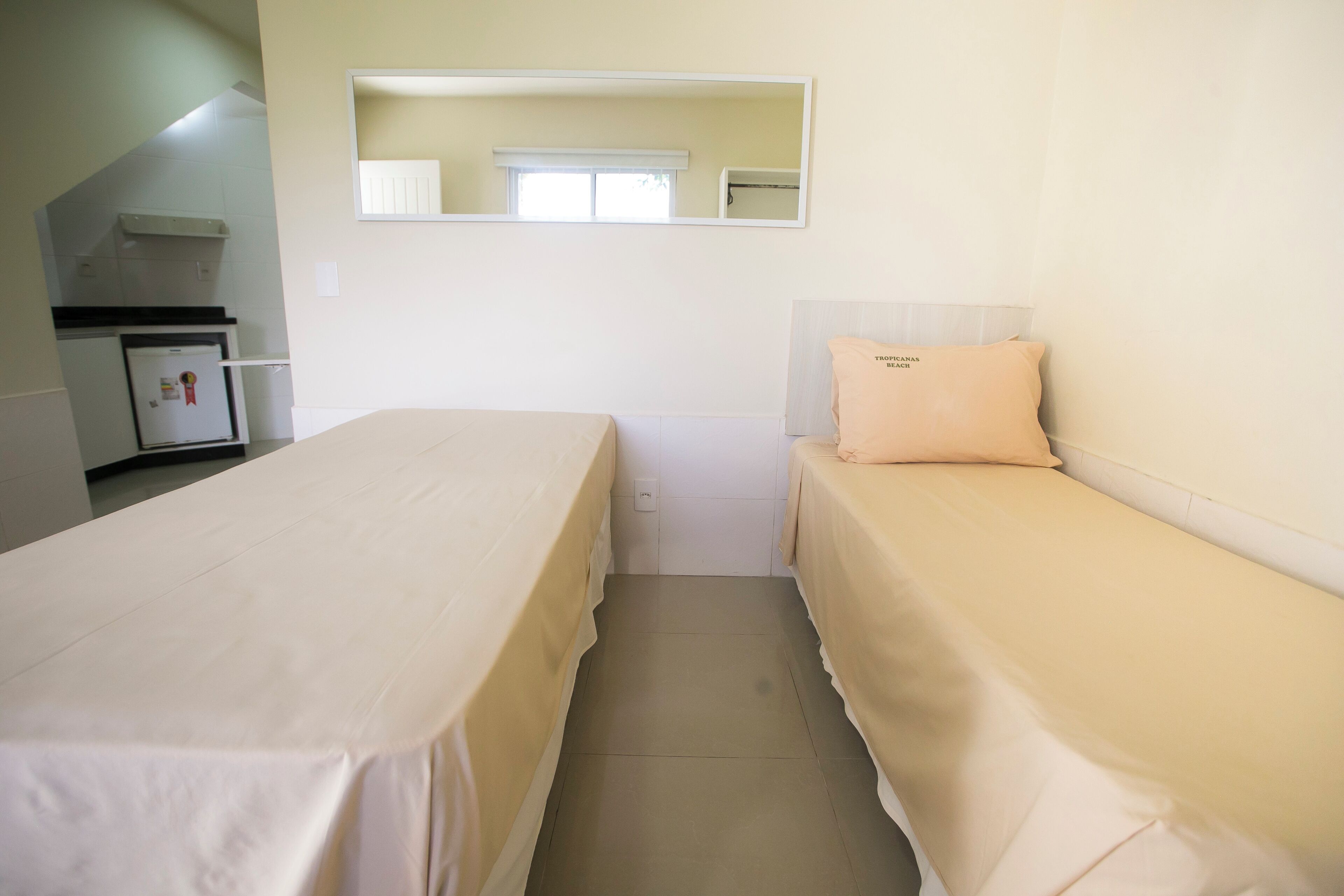 A bedroom with two beds, a small kitchenette, a mirror, and a window.