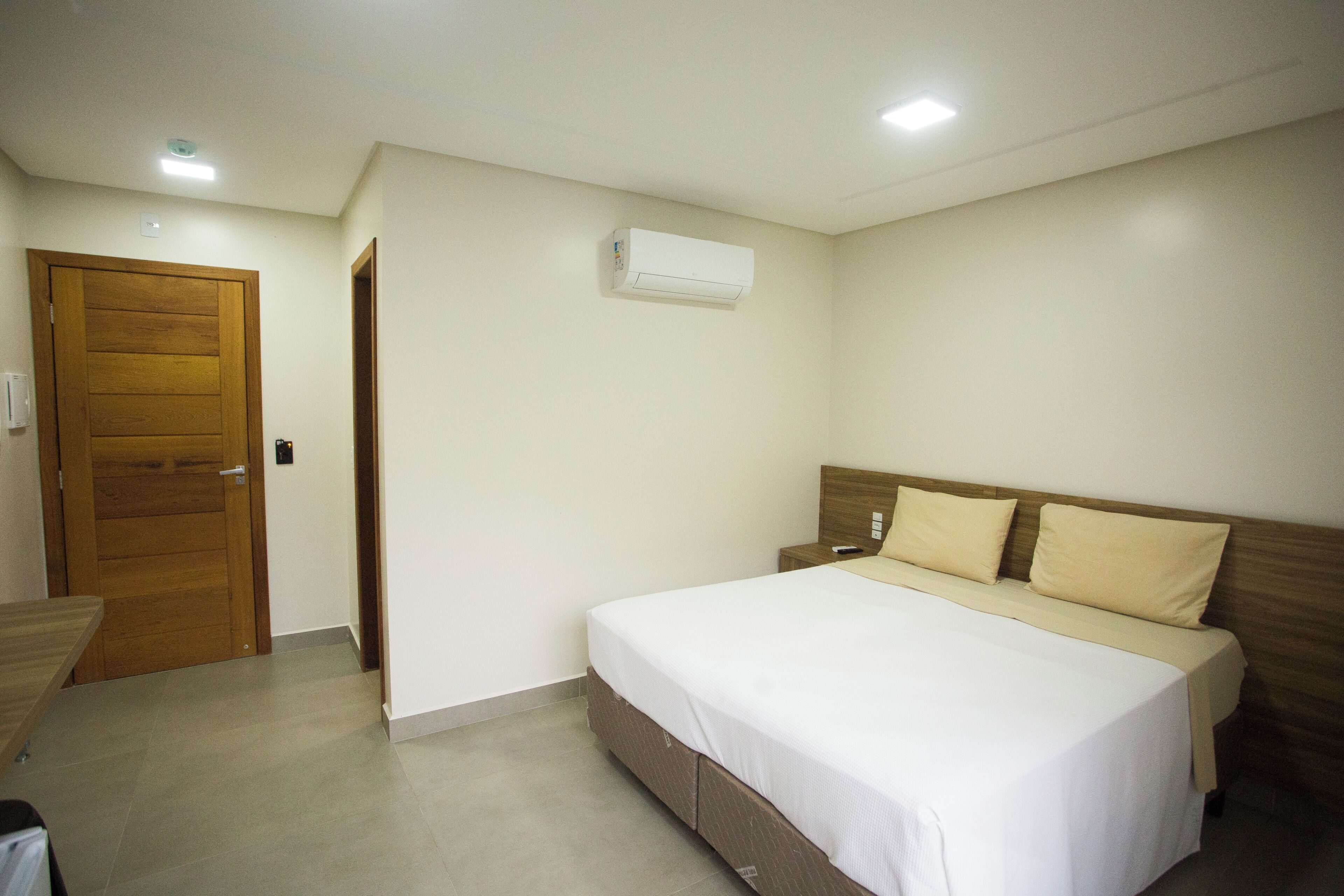 A bedroom with a bed, a wooden door, a wall-mounted air conditioner, and a bedside table.
