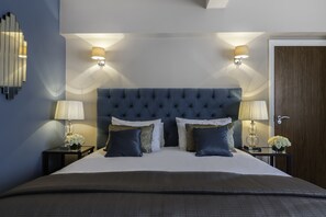 A neatly made bed with a tufted headboard, two bedside lamps, and a nightstand with a vase of flowers. - Arlington House (London)
