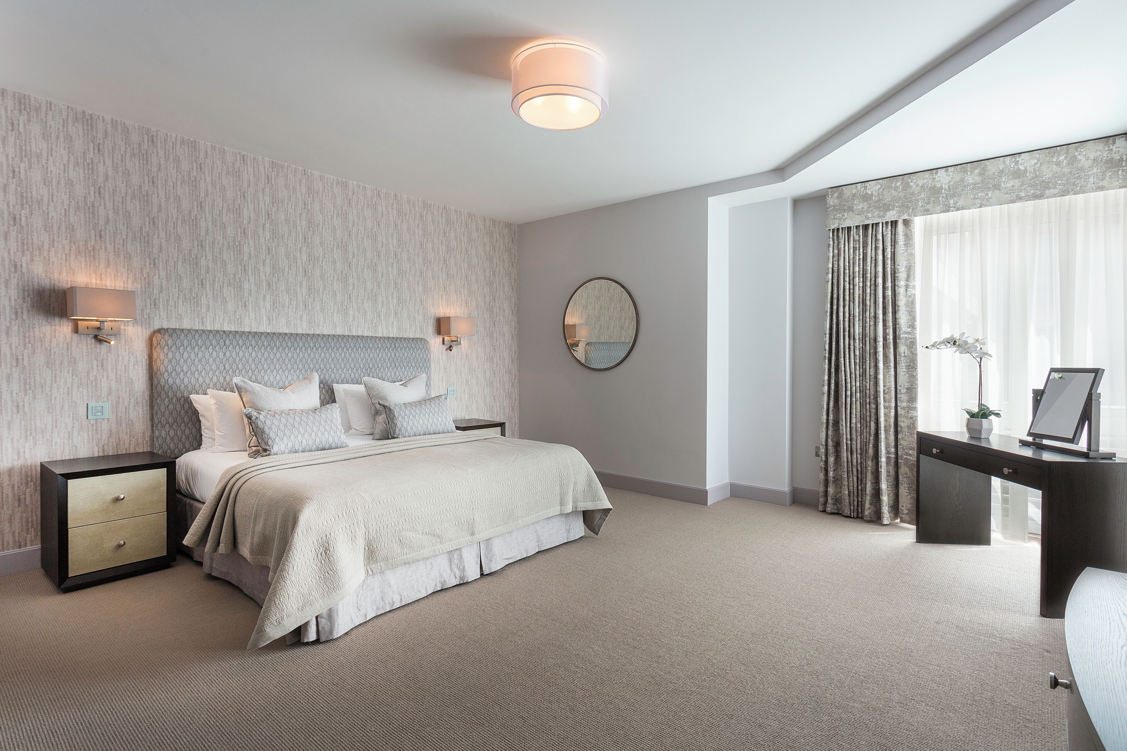 A bedroom with a bed, bedside tables, a desk, a mirror, and a large window with curtains.