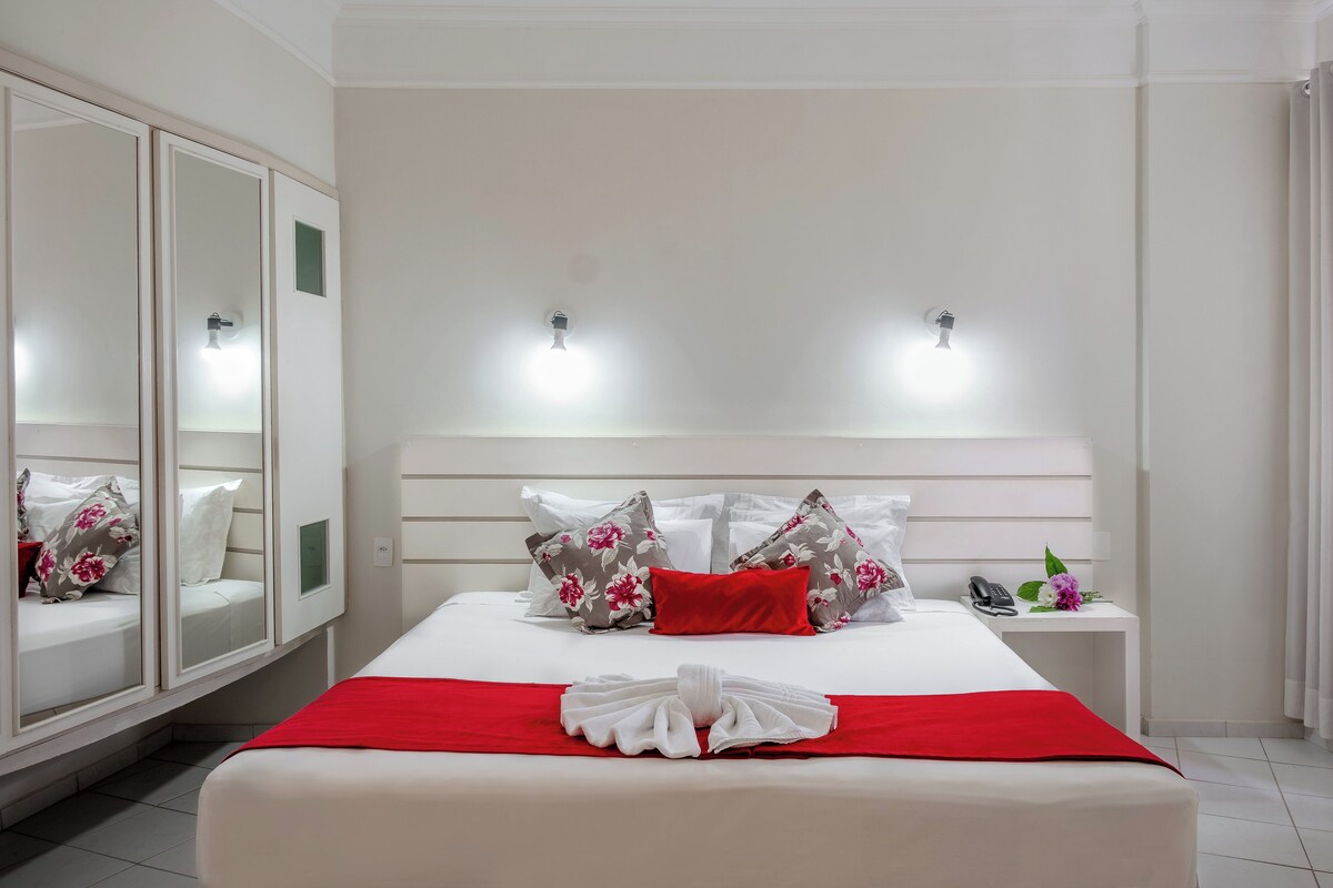 A neatly made bed with a red blanket and a towel, a bedside phone, and a small table with a flower.
