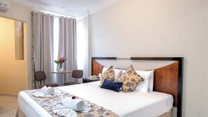 Une chambre d’hôtel avec un grand lit, un bureau avec une chaise, une petite table avec un vase de fleurs et une fenêtre avec des rideaux.