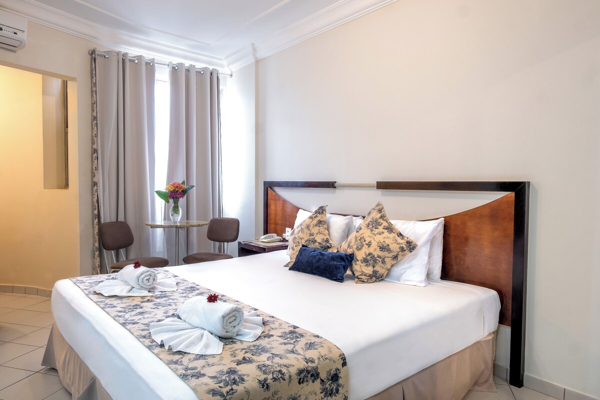 A hotel room with a large bed, a desk with a chair, a small table with a vase of flowers, and a window with curtains.